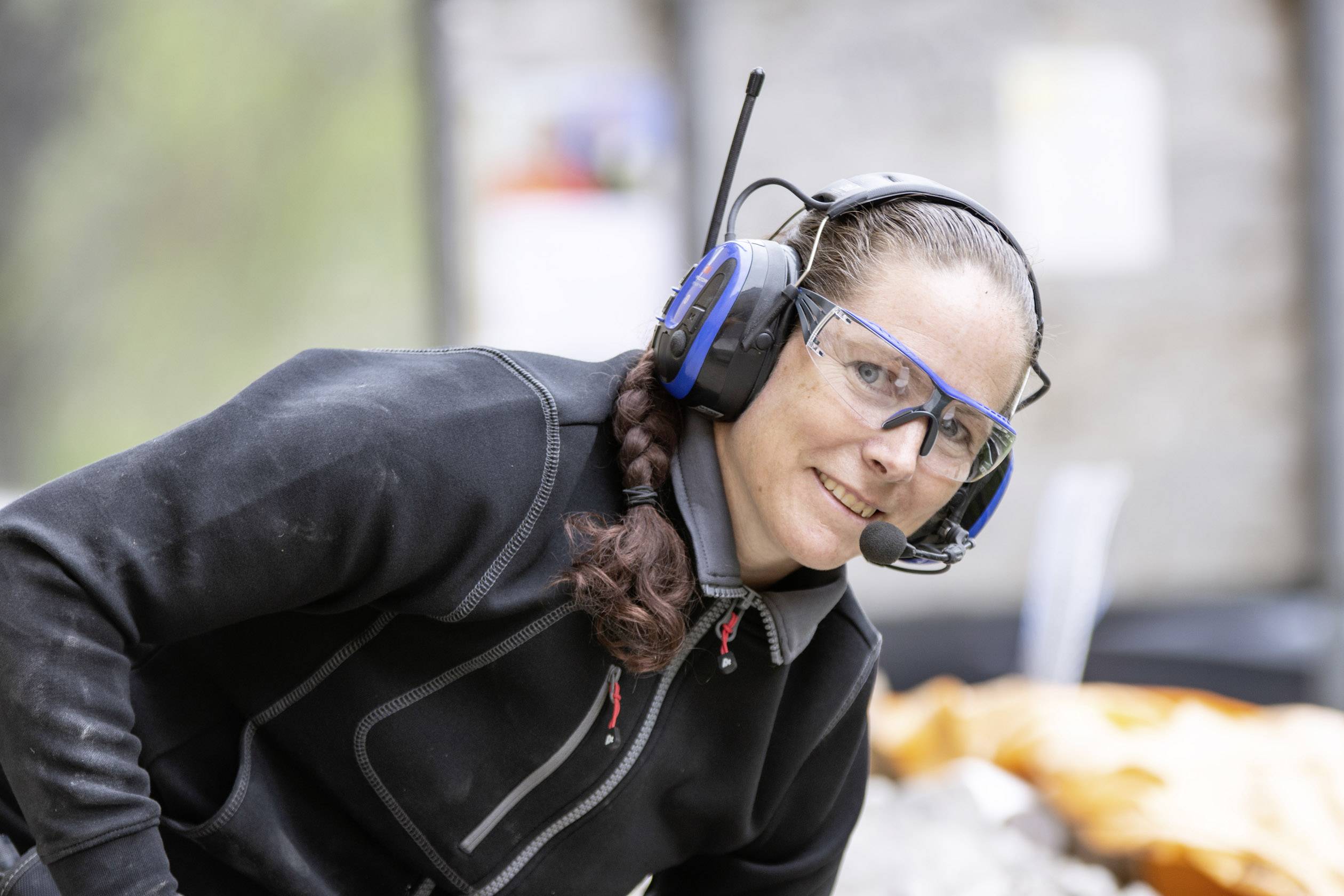 Eine Person mit Schutzbrille und Headset lächelt, während sie draußen arbeitet.