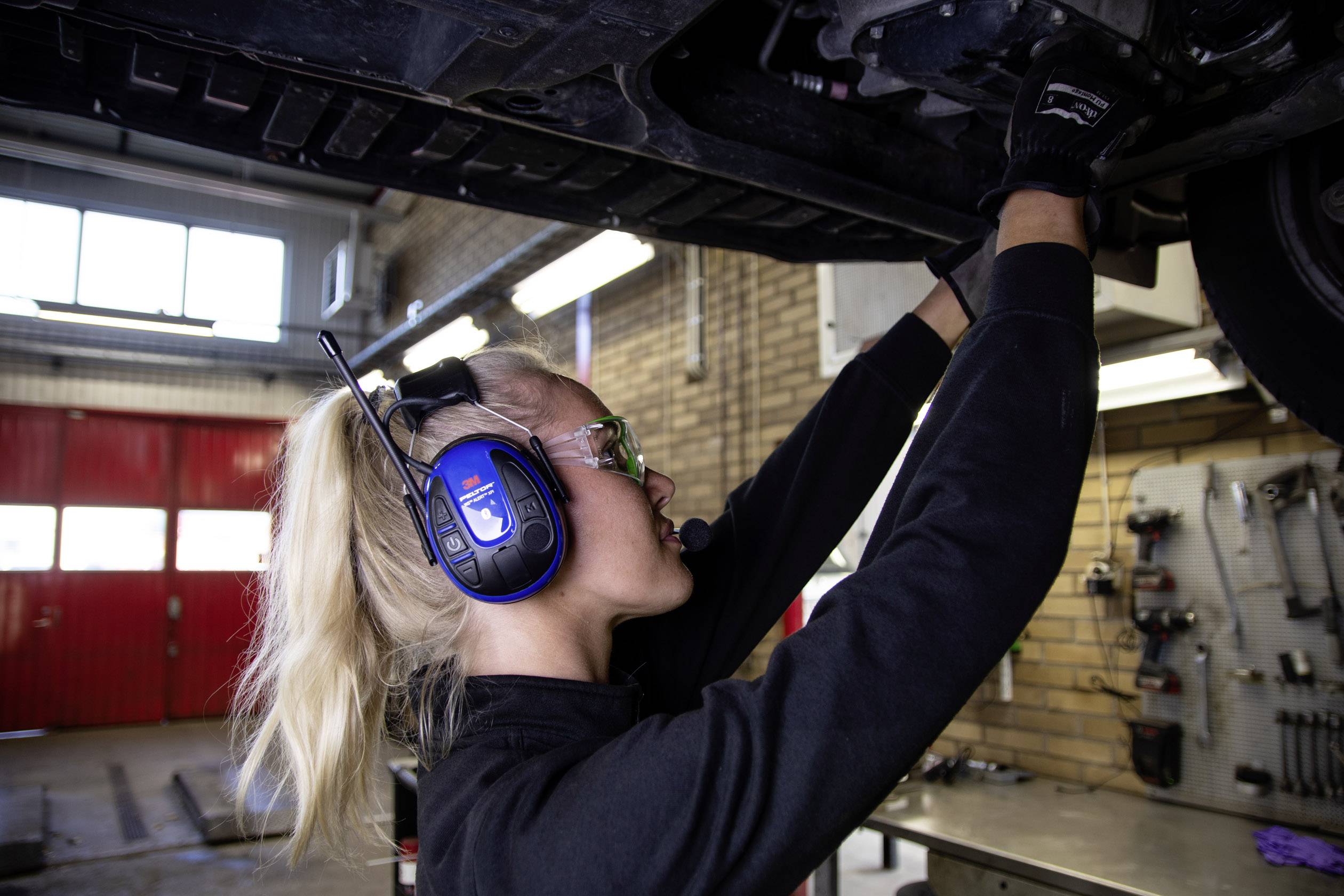 Eine Person mit Schutzbrille und Kopfhörern arbeitet in einer Werkstatt unter einem angehobenen Fahrzeug. Handwerkliche Tätigkeit im Vordergrund.