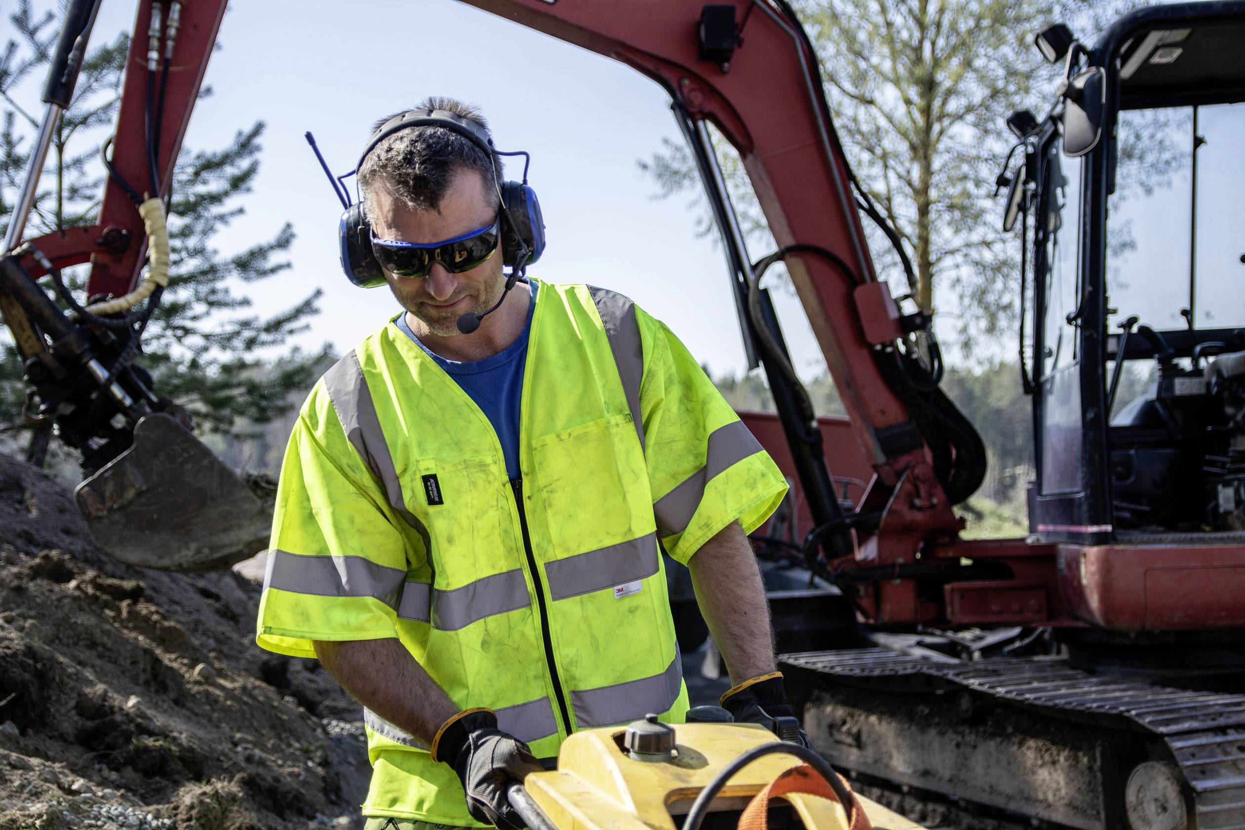 Ein Bauarbeiter in gelber Warnkleidung arbeitet mit einem Werkzeug vor einem Bagger auf einer Baustelle im Freien.