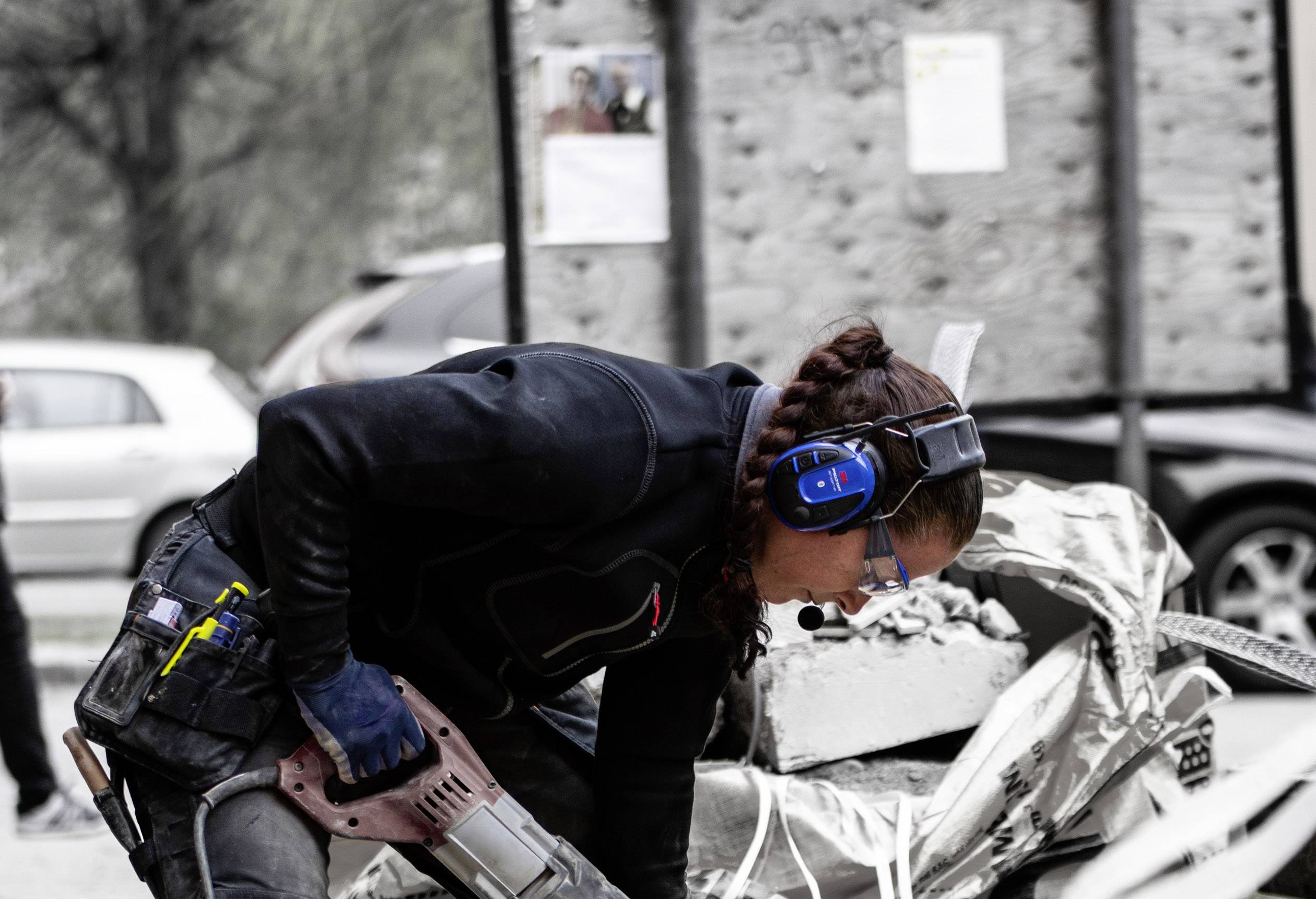 Eine Person bohrt konzentriert mit einem großen Bohrer in einem Baustellenbereich, trägt Gehörschutz und Arbeitskleidung. Im Hintergrund unscharfe Autos.