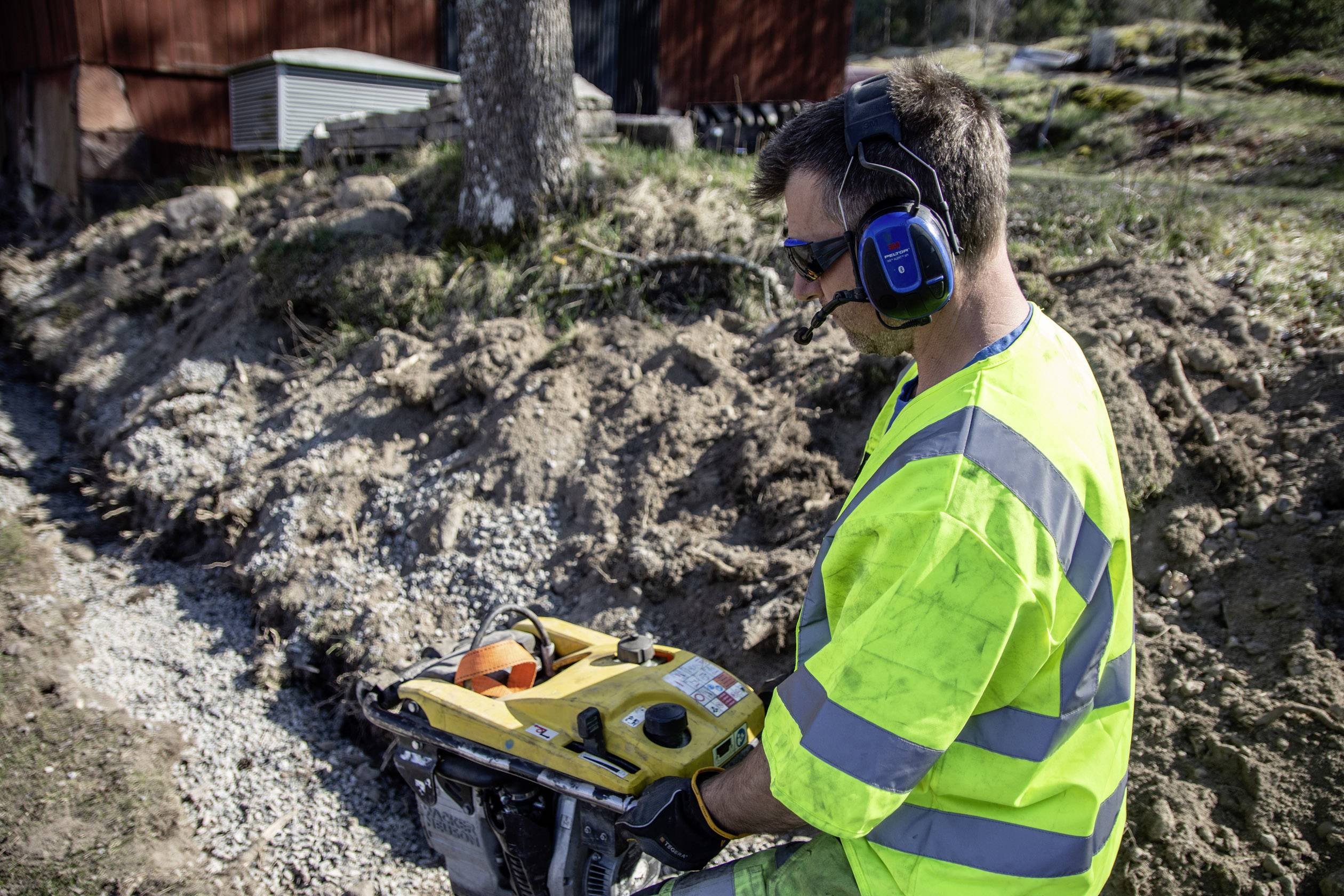 Ein Arbeiter in gelber Schutzkleidung und Gehörschutz bedient eine Baumaschine im Freien, um Erde und Kies auf einem Grundstück zu bearbeiten.