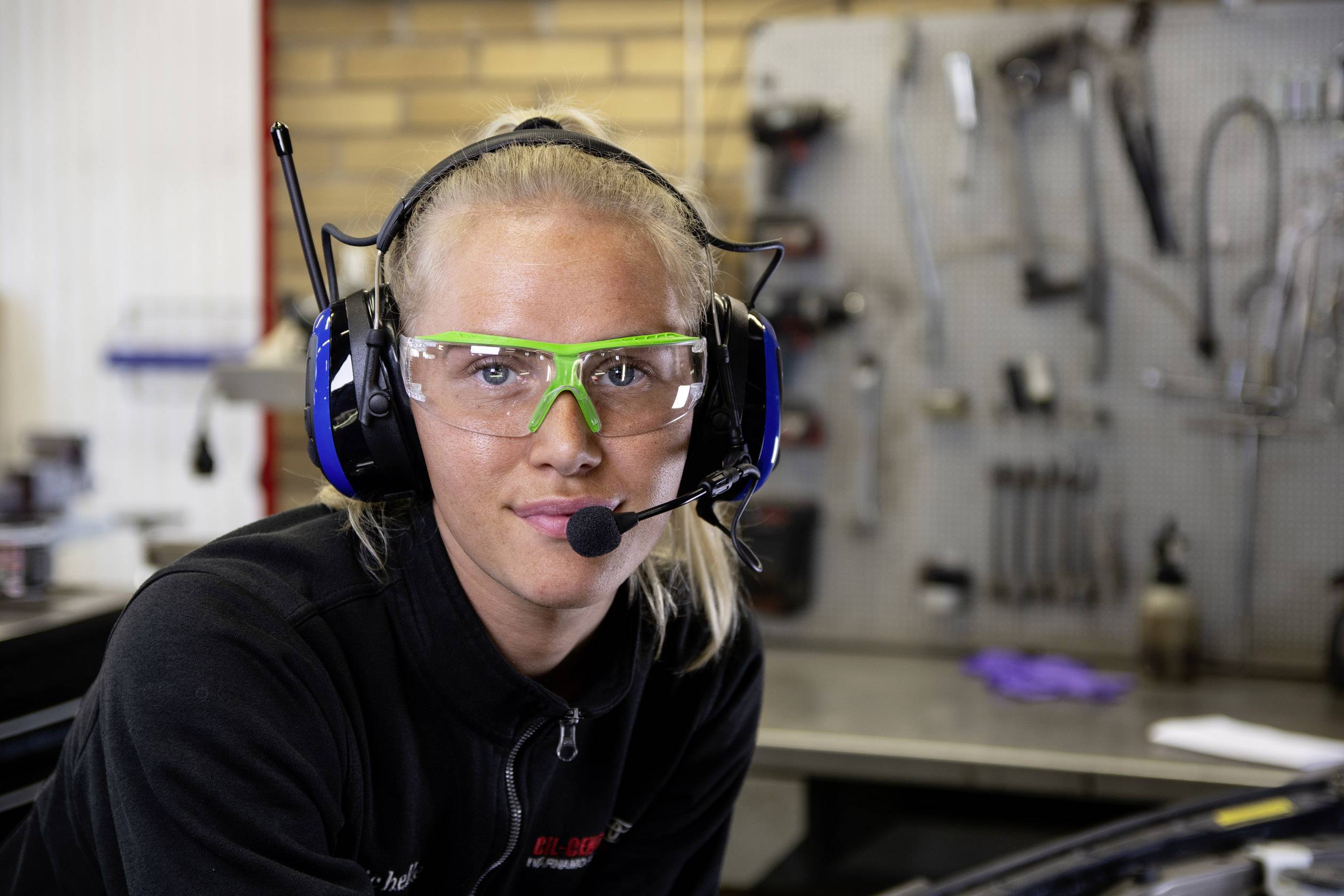 Eine Frau in Schutzbrille und Kopfhörer mit Mikrofon in einer Werkstattumgebung. Im Hintergrund sind Werkzeuge sichtbar.