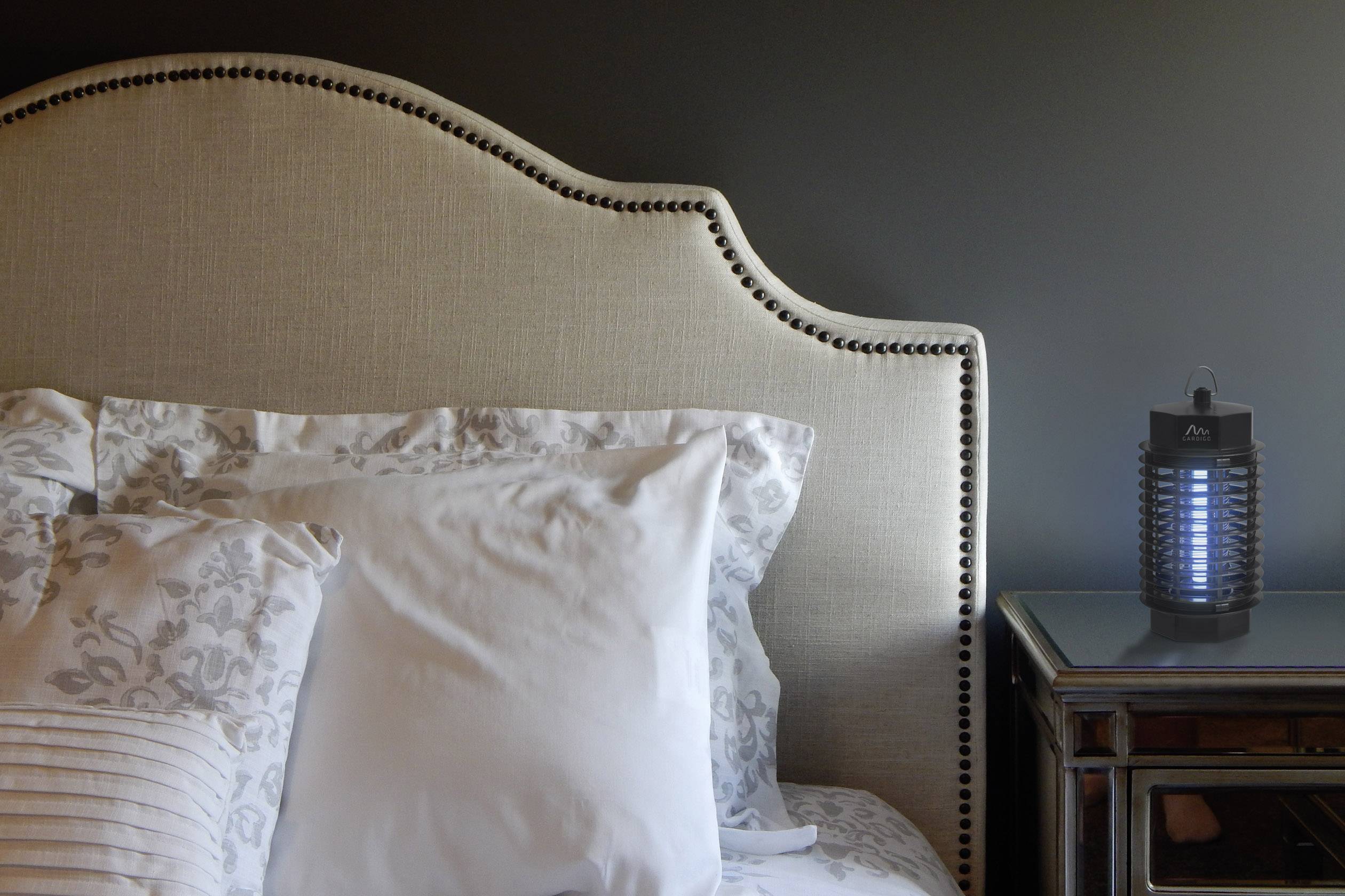 Ein Bett mit weißen Kissen steht in einem Schlafzimmer. Auf dem Nachttisch rechts steht eine elektrische Insektenlampe.