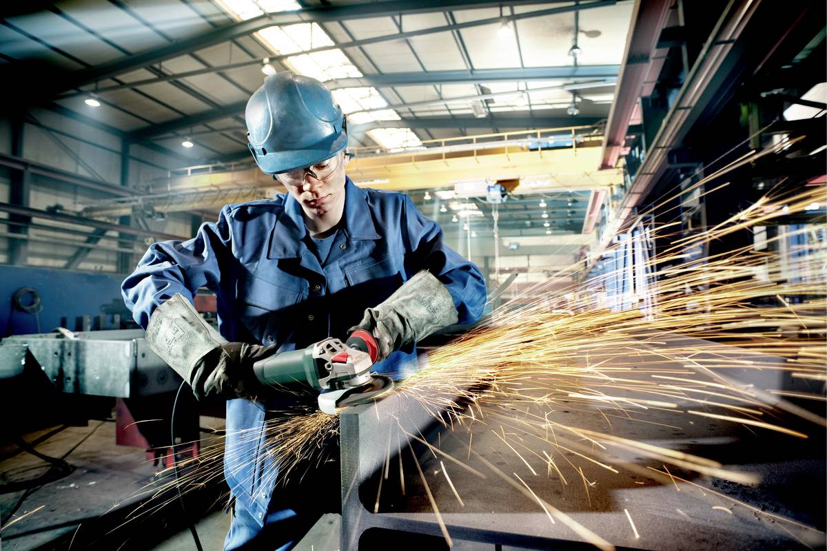 Ein Arbeiter in Schutzkleidung schleift Metall mit einem Winkelschleifer in einer Fabrikhalle. Funken fliegen in alle Richtungen.