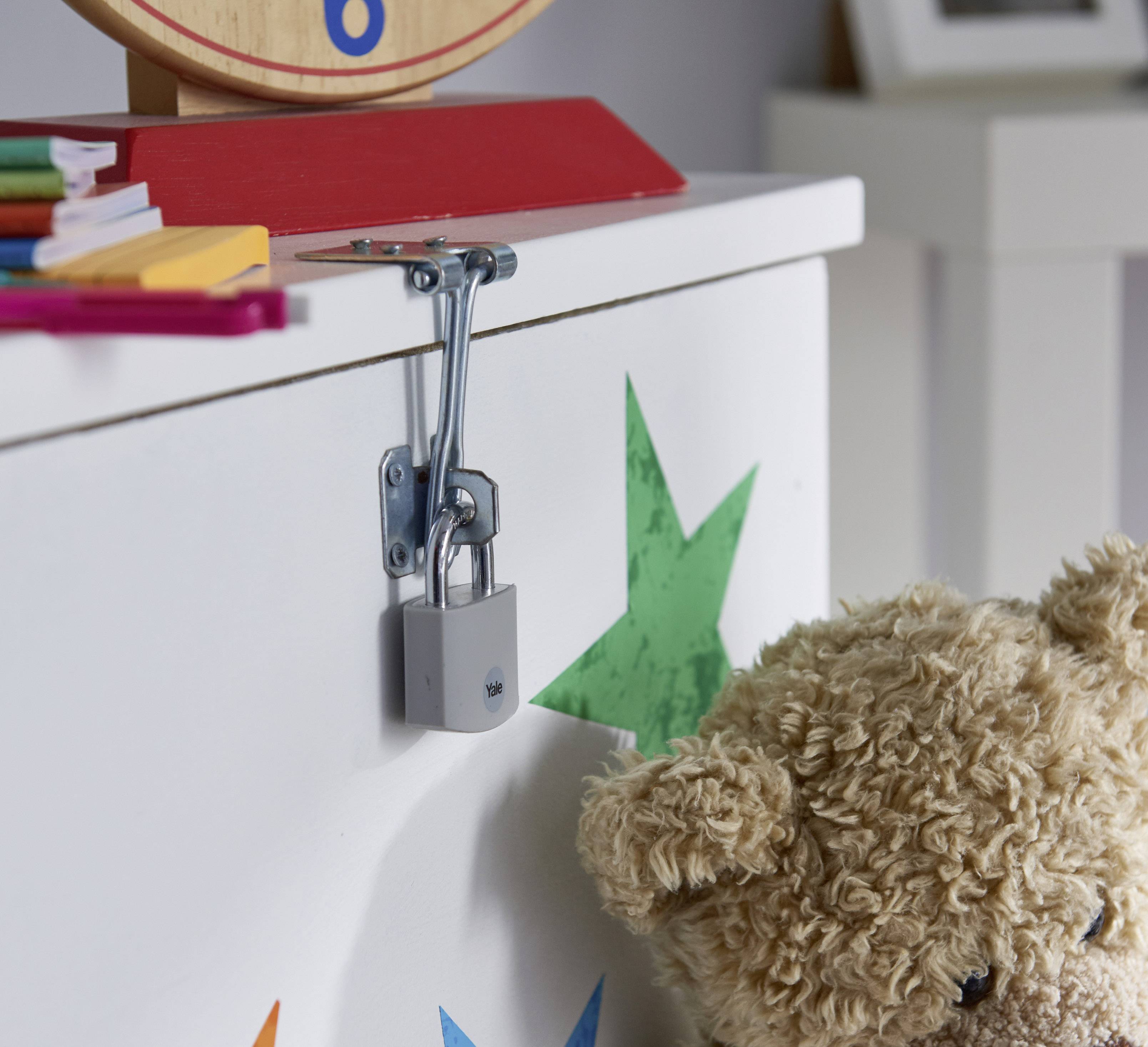 Ein Kinderzimmer mit bunter Spieltruhe, gesichert mit einem Schloss, und einem Plüschbär im Vordergrund. Bürobedarf im Hintergrund.