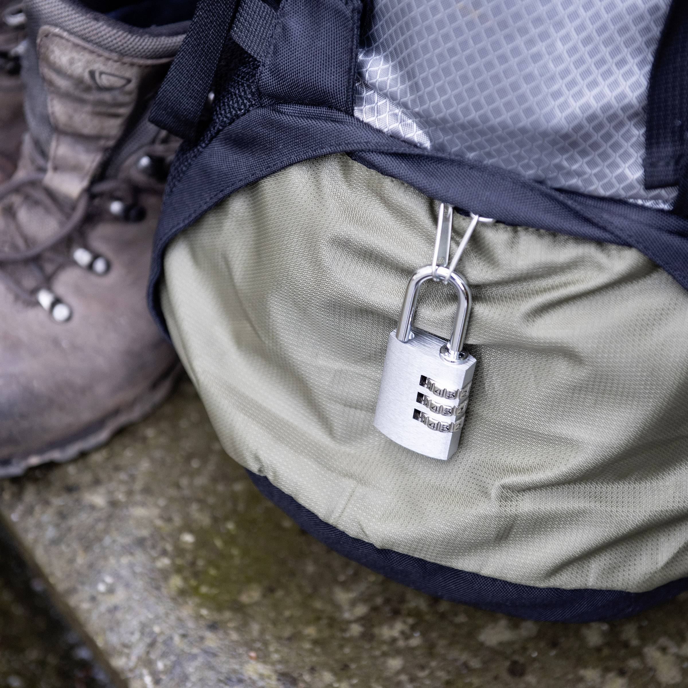 'Einem Rucksack ist ein Vorhängeschloss befestigt. Daneben steht ein Wanderschuh auf einem steinigen Boden.'