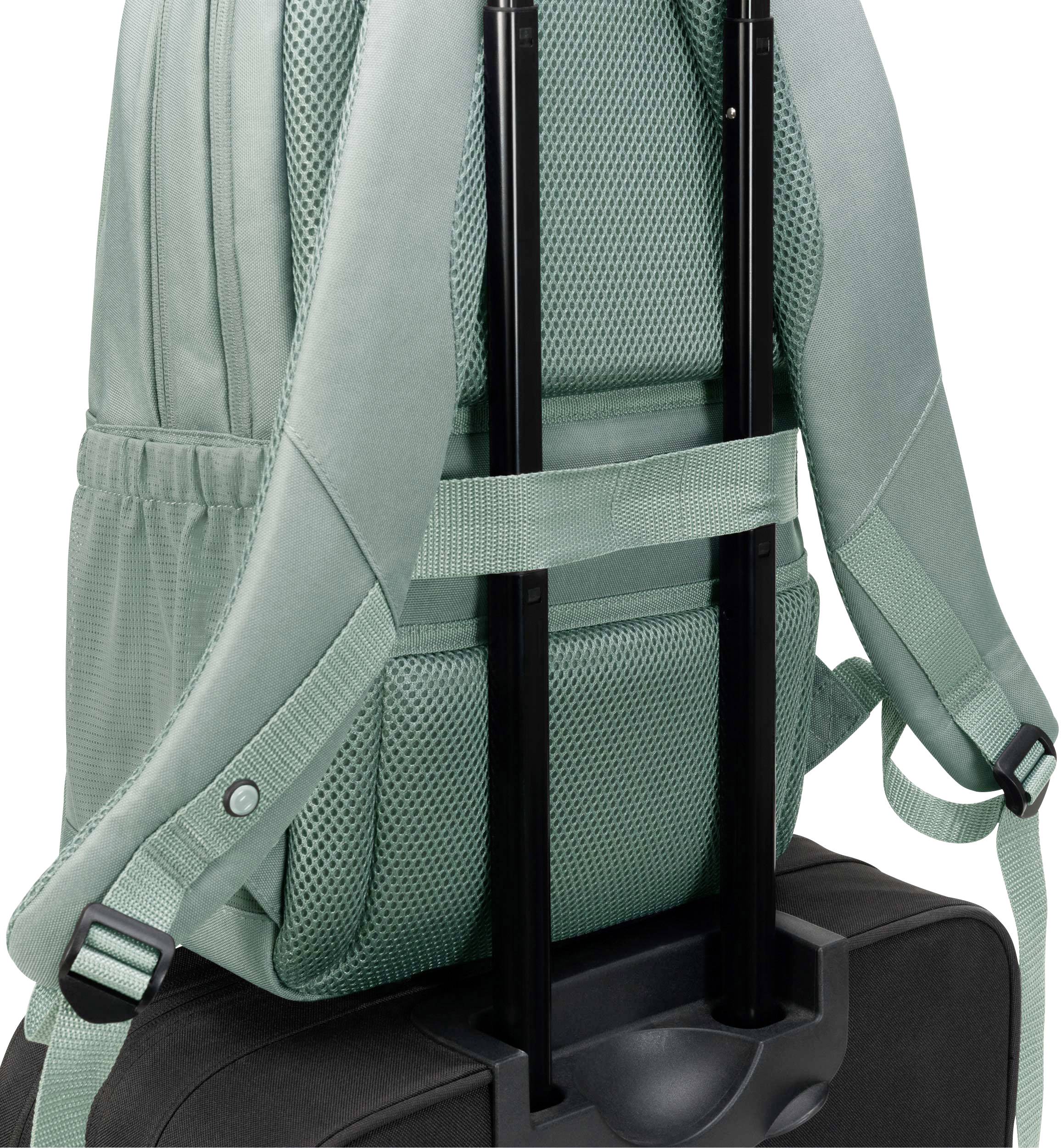 Ein grüner Rucksack ist auf einem schwarzen Rollkoffer befestigt, indem er mit einem Gurt über die Teleskopgriffe gesichert ist.