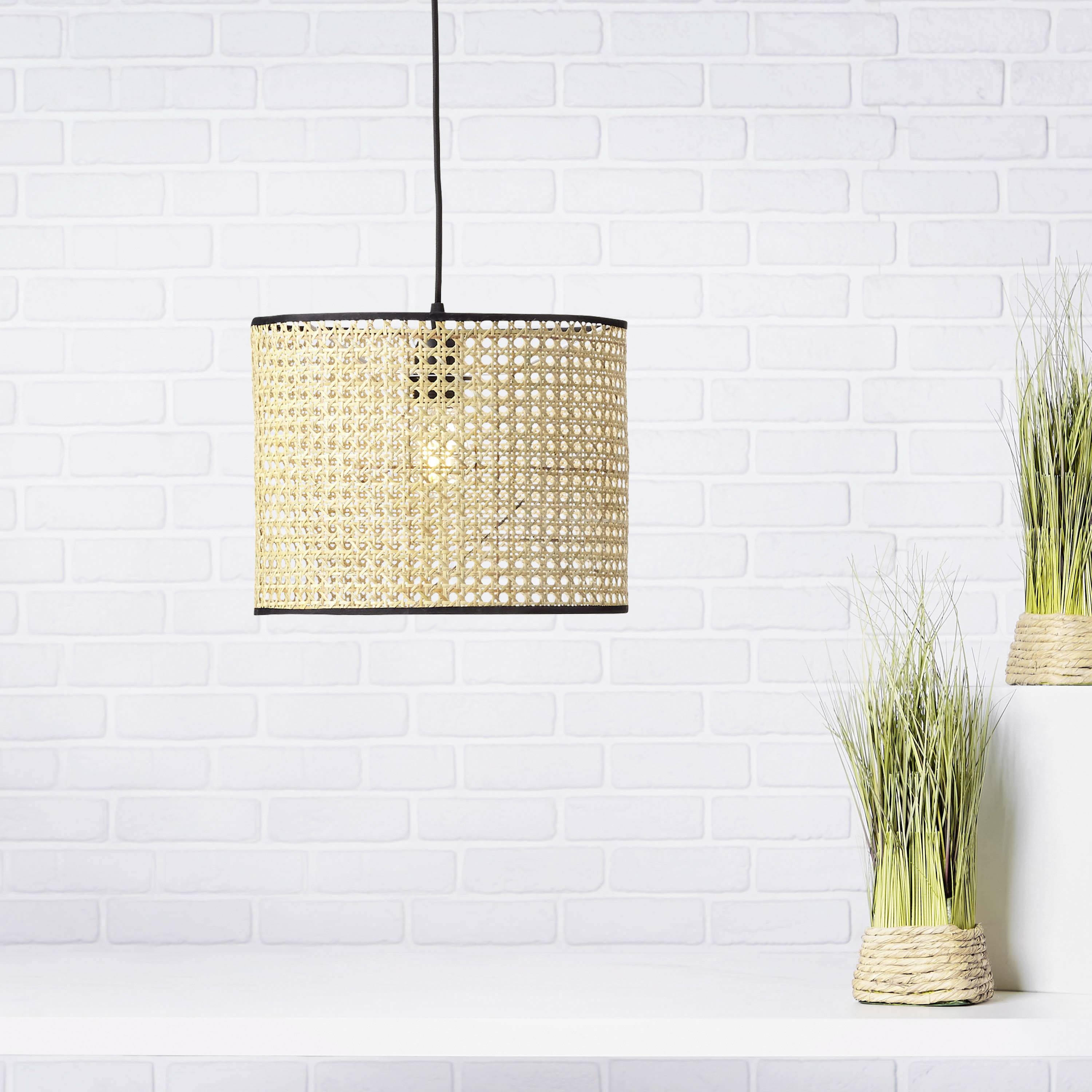 Eine hängende Lampe mit geflochtenem Schirm vor einer weißen Ziegelwand. Zwei Dekopflanzen stehen rechts auf einem Tisch.