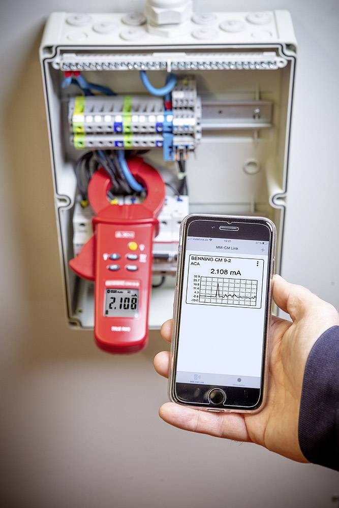 Eine Person hält ein Smartphone, das eine Messung von 2108 mA von einem Gerät in einem geöffneten Verteilerkasten anzeigt.