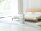 Luftreiniger steht in einem minimalistisch eingerichteten Schlafzimmer neben einem Bett mit Holzrahmen und einer weißen Lampe.