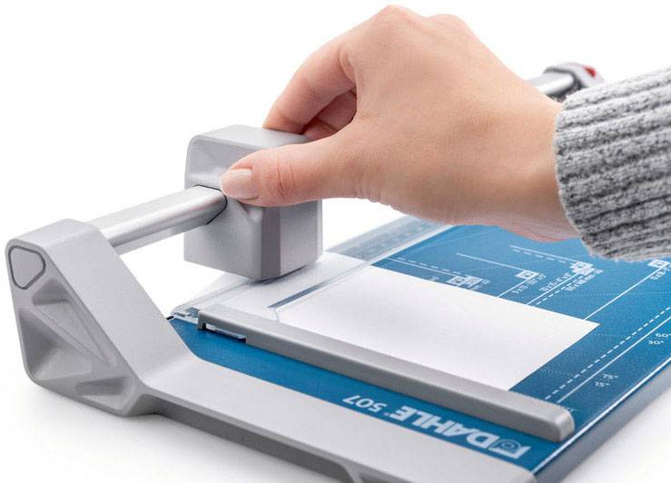 Eine Hand bedient einen Papierschneider, um ein Blatt Papier präzise zu schneiden. Der Schneider hat ein blaues Lineal mit Maßen.