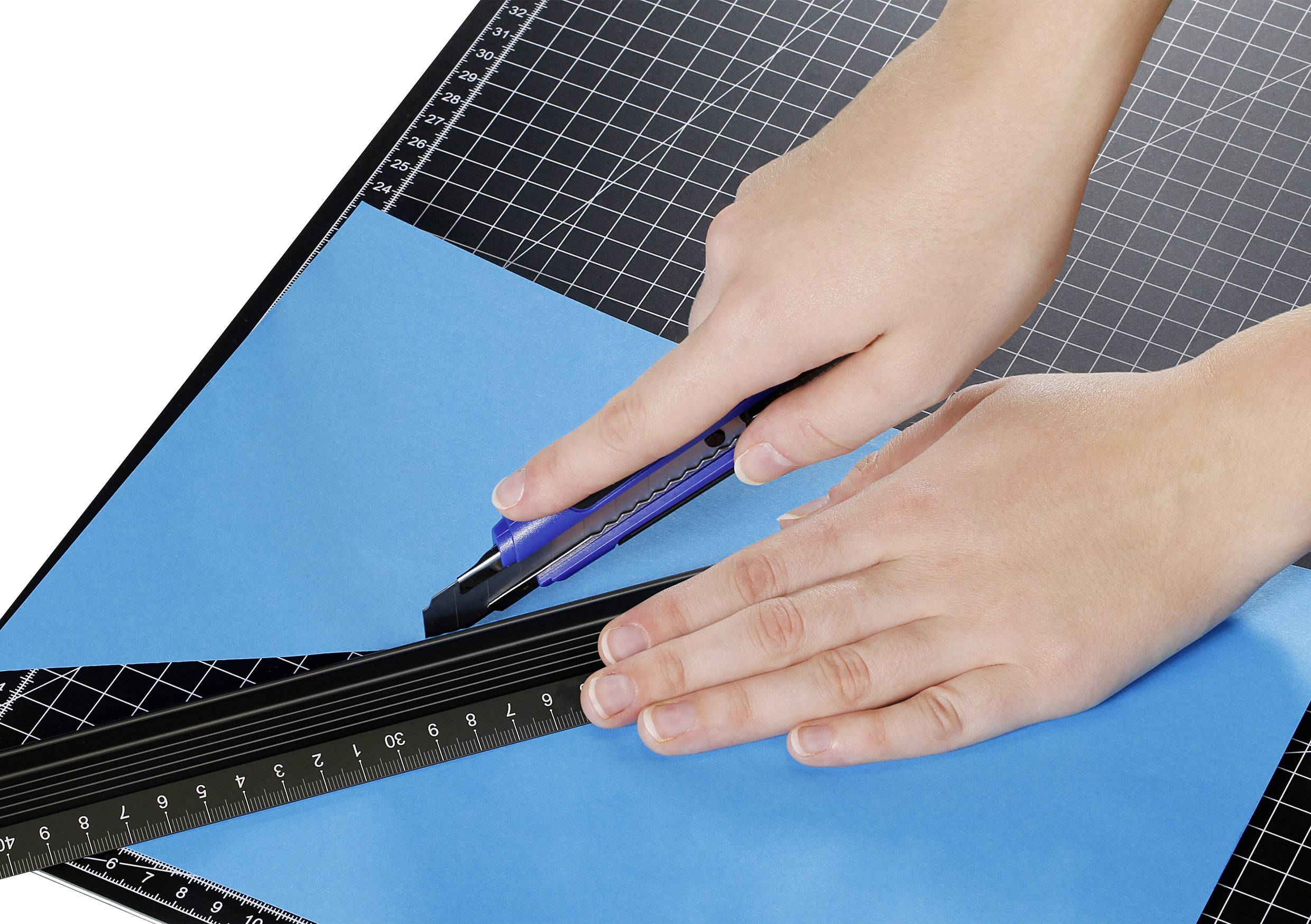 Hände schneiden blaues Papier mit einem Cuttermesser und Lineal auf einer Schneidematte.