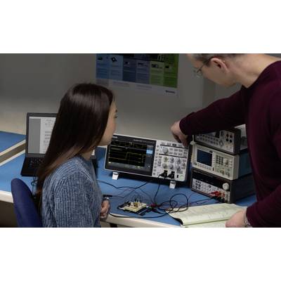 Ein Mann zeigt einer Frau ein Oszilloskop in einem Labor. Sie analysieren elektronische Schaltungen auf einem Tisch mit Laptops.