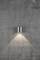 Eine einzelne silberne Wandlampe strahlt warmes Licht auf eine graue Betonwand und erzeugt einen weichen Lichtkegel nach unten.