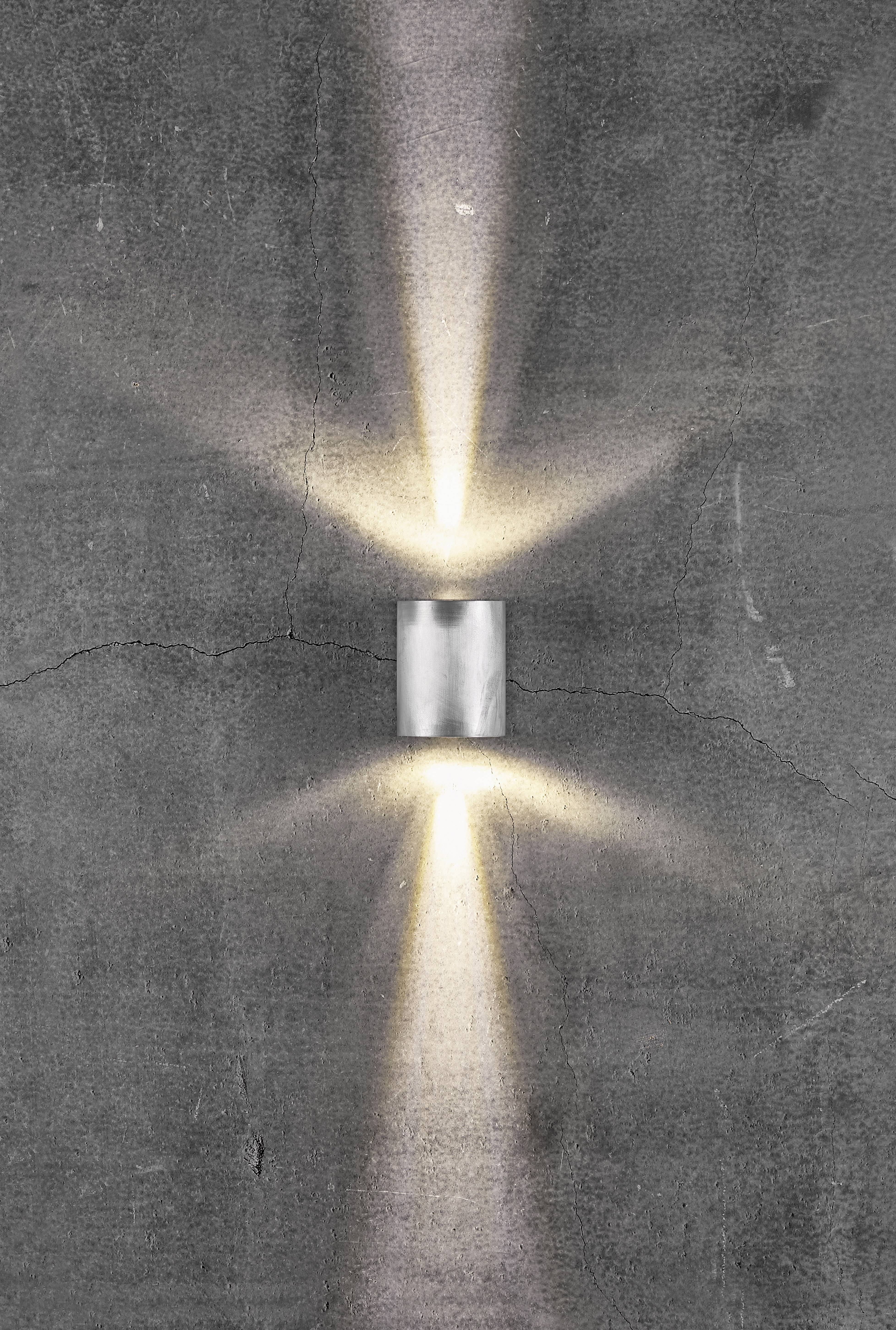 Eine glänzende Wandleuchte strahlt Licht nach oben und unten auf eine graue, leicht rissige Oberfläche.