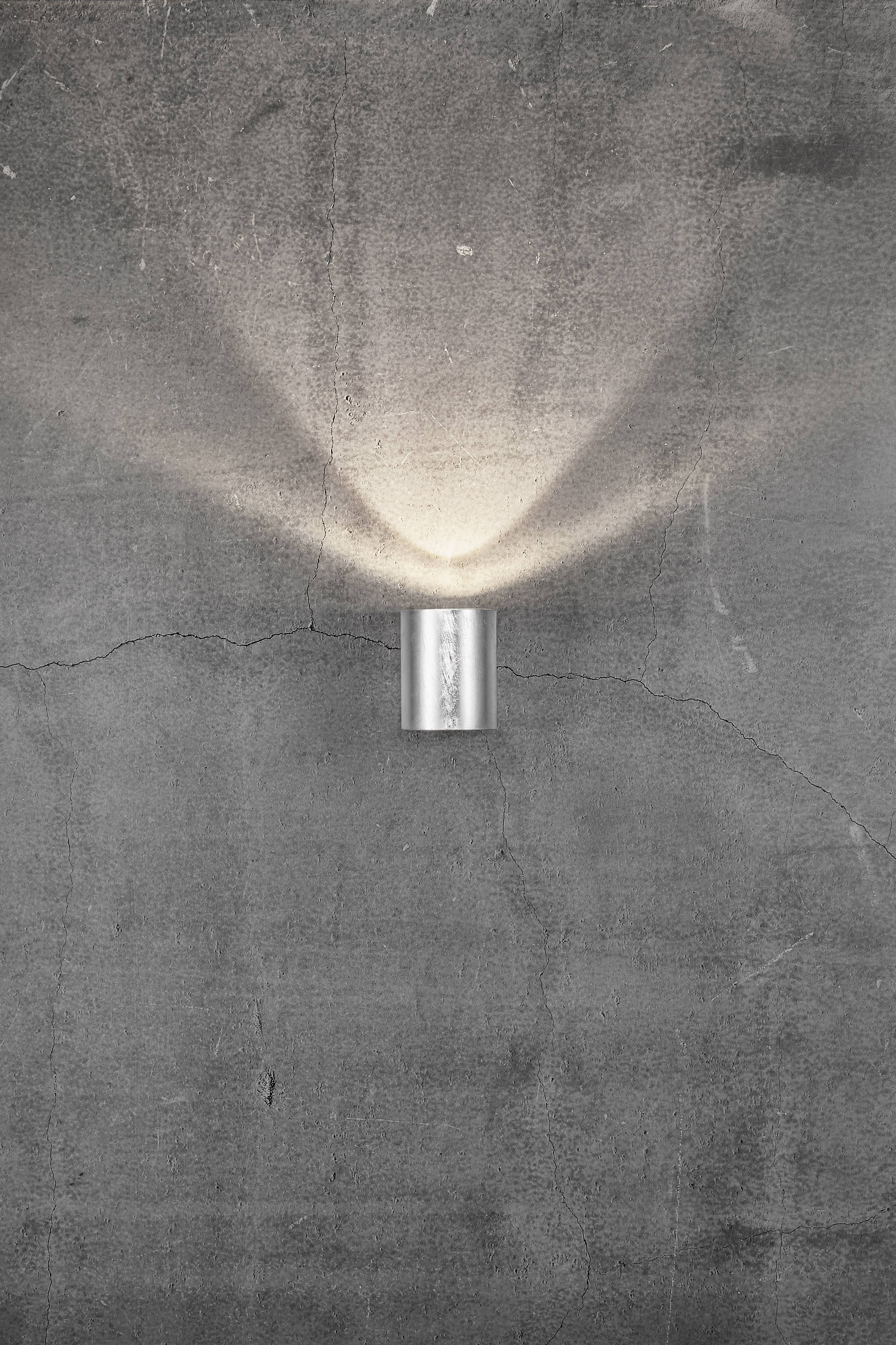 Wandlampe aus Metall in moderner Form, wirft warmes Licht nach oben und unten auf eine graue, leicht rissige Betonwand.