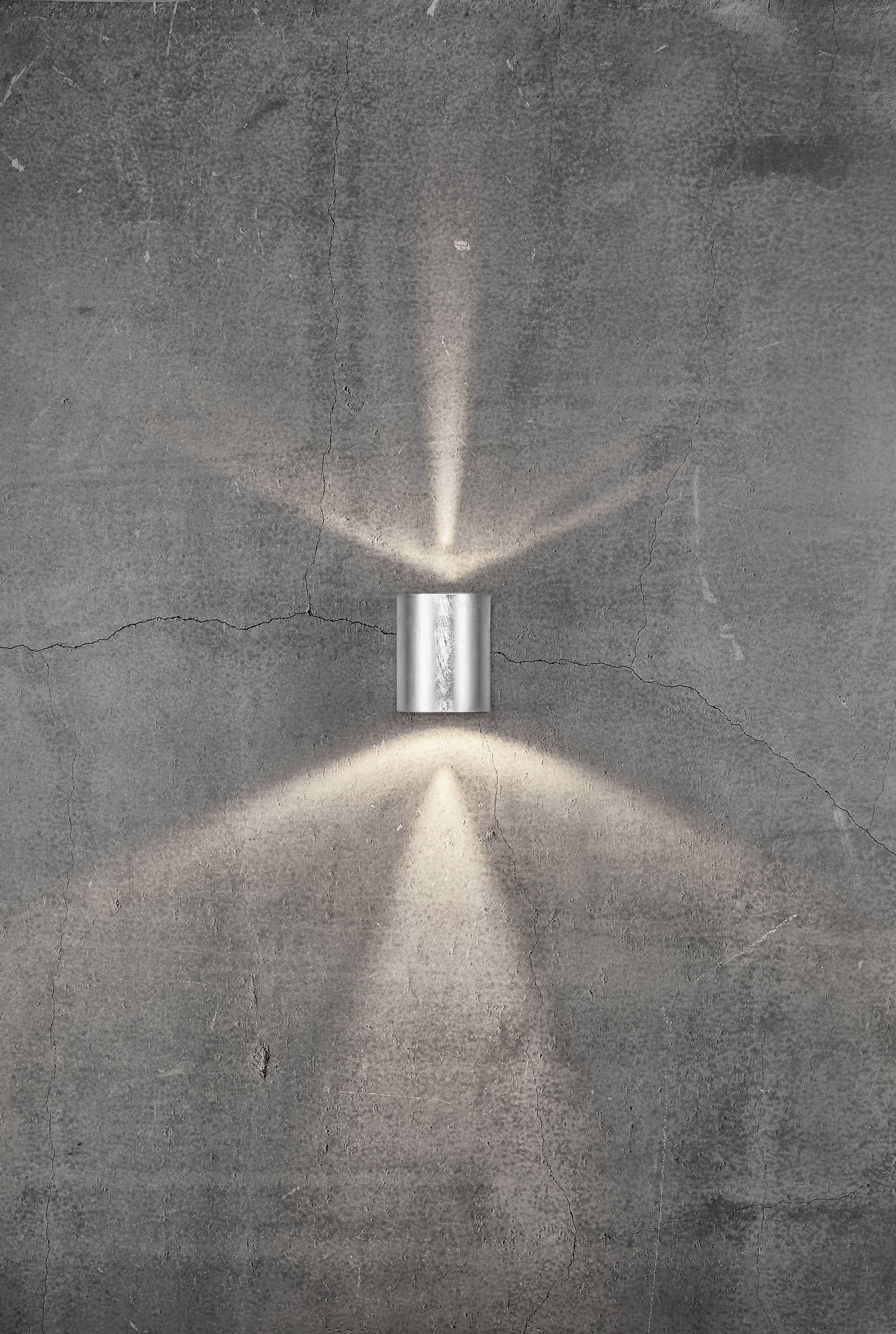 Eine zylindrische Wandleuchte strahlt Lichtstrahlen nach oben und unten auf eine graue, strukturierte Betonwand.