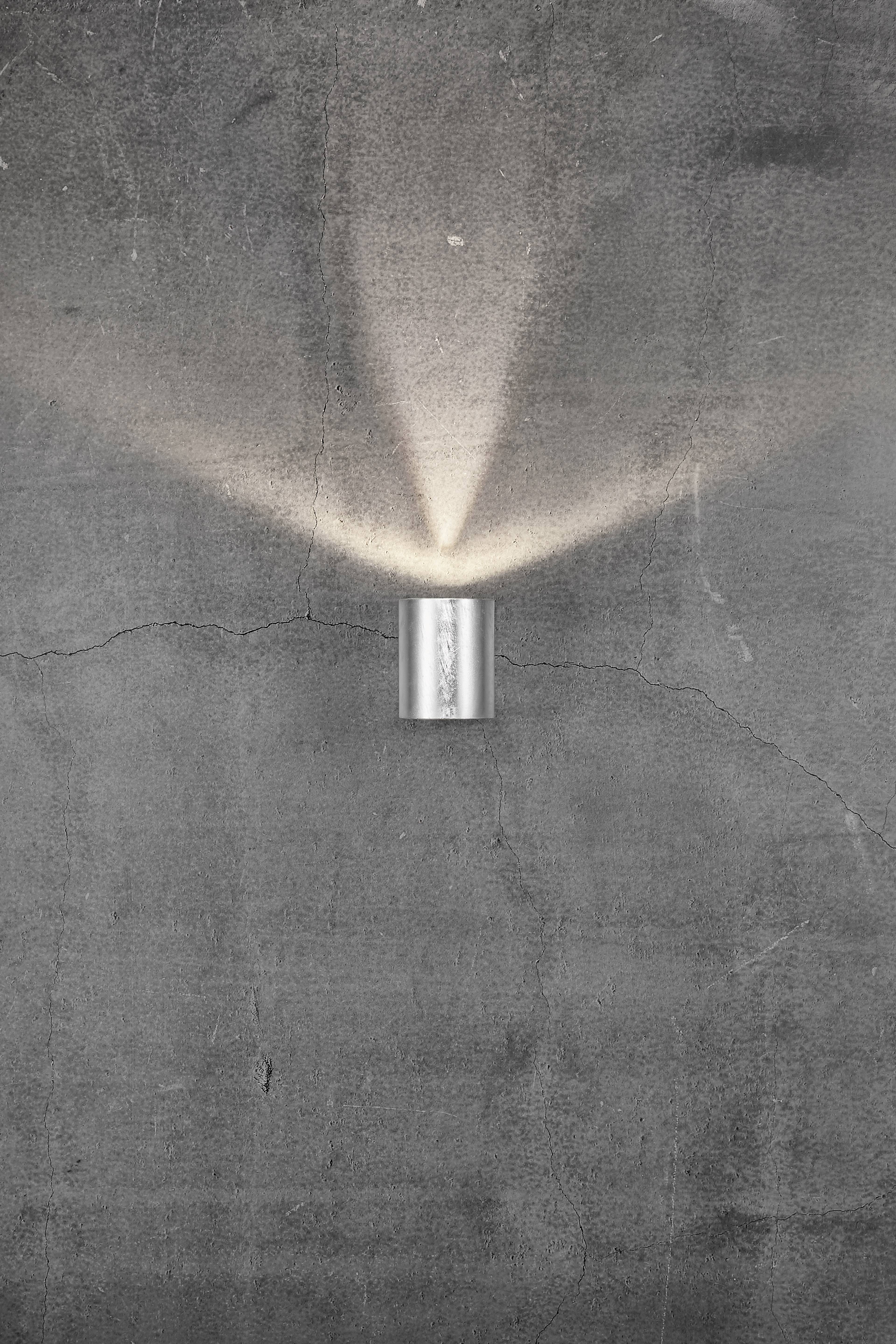 Eine silberne Wandlampe an einer grauen Betonwand leuchtet nach oben und unten, erzeugt ein kreuzförmiges Lichtmuster.