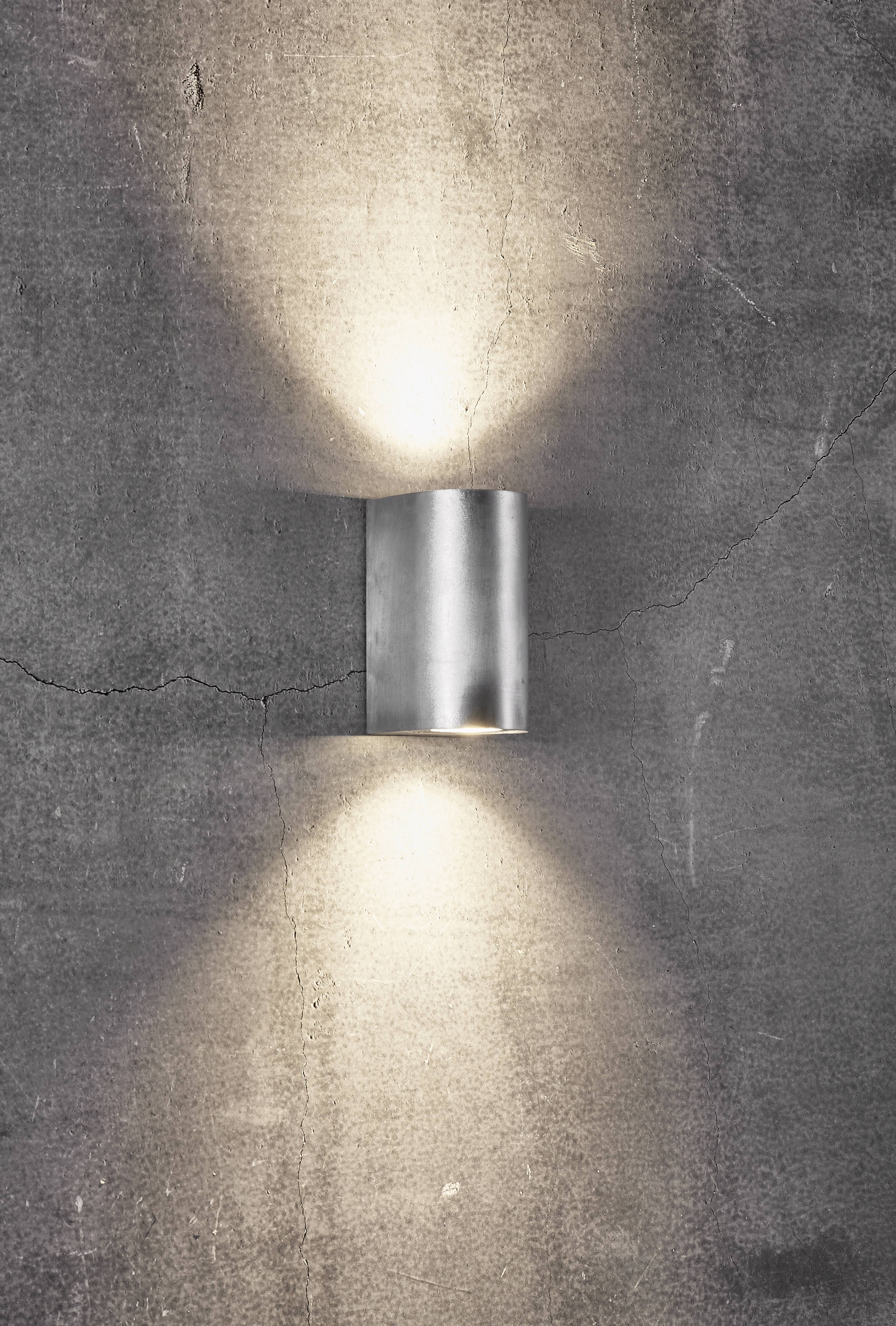Eine zylindrische Wandleuchte aus poliertem Metall an einer grauen, strukturierten Wand beleuchtet nach oben und unten.
