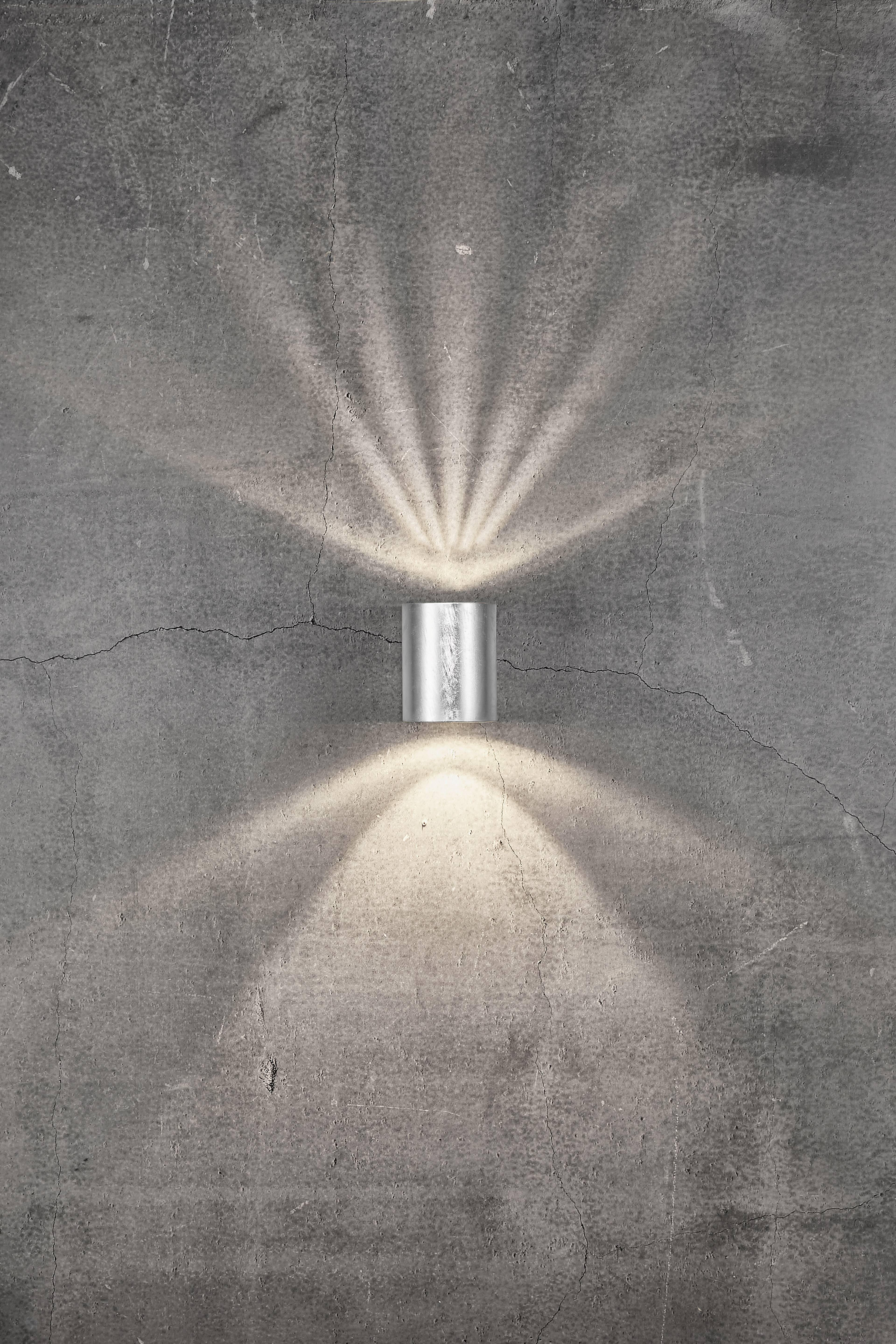 Eine Metalllampe an einer grauen Betonwand strahlt Lichtstrahlen nach oben und unten, wodurch ein atmosphärisches Muster entsteht.