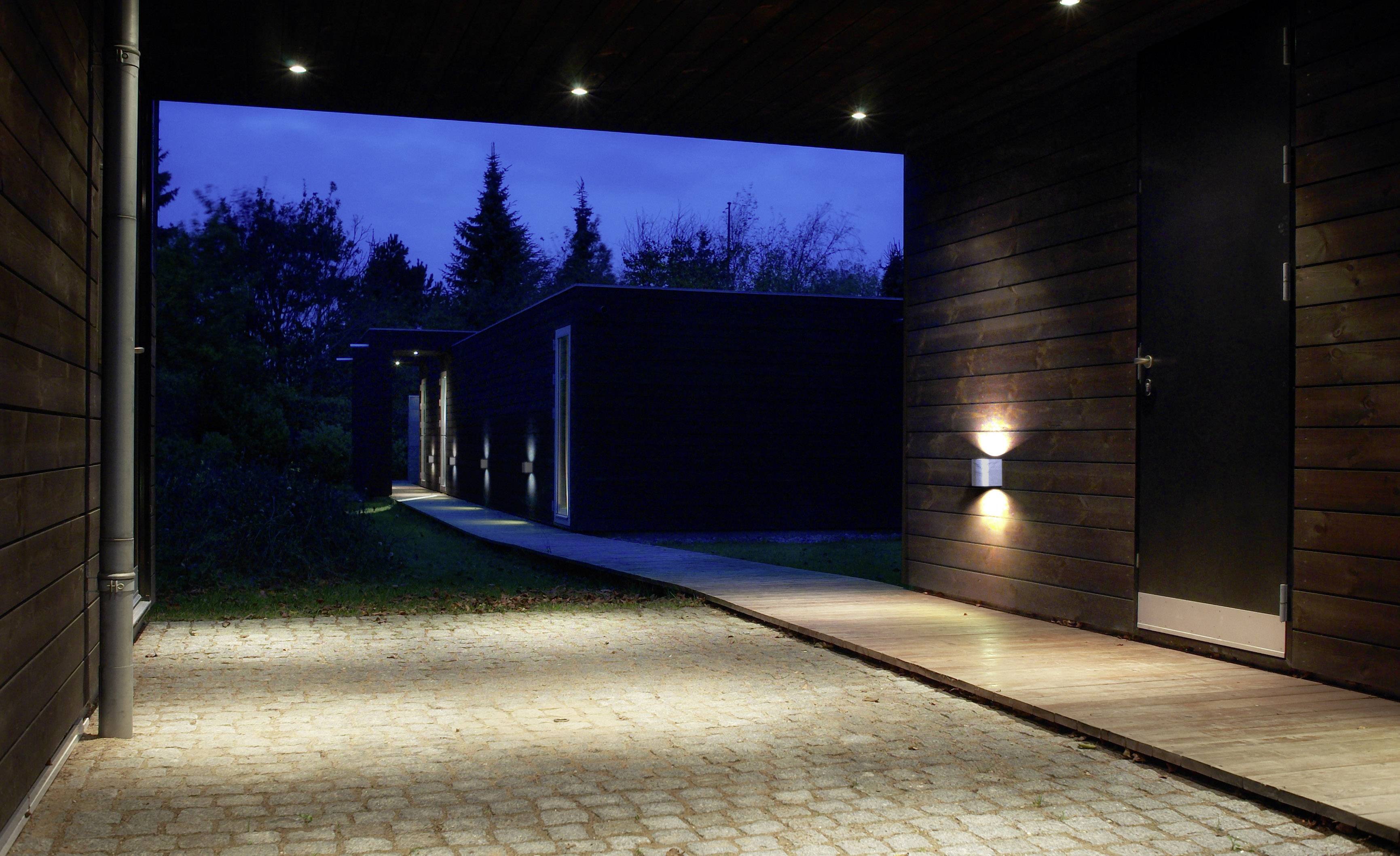 Ein moderner, beleuchteter Eingang mit Wegführung in einem Wohnbereich bei Nacht. Rechteckige Architektur, umgeben von Bäumen.