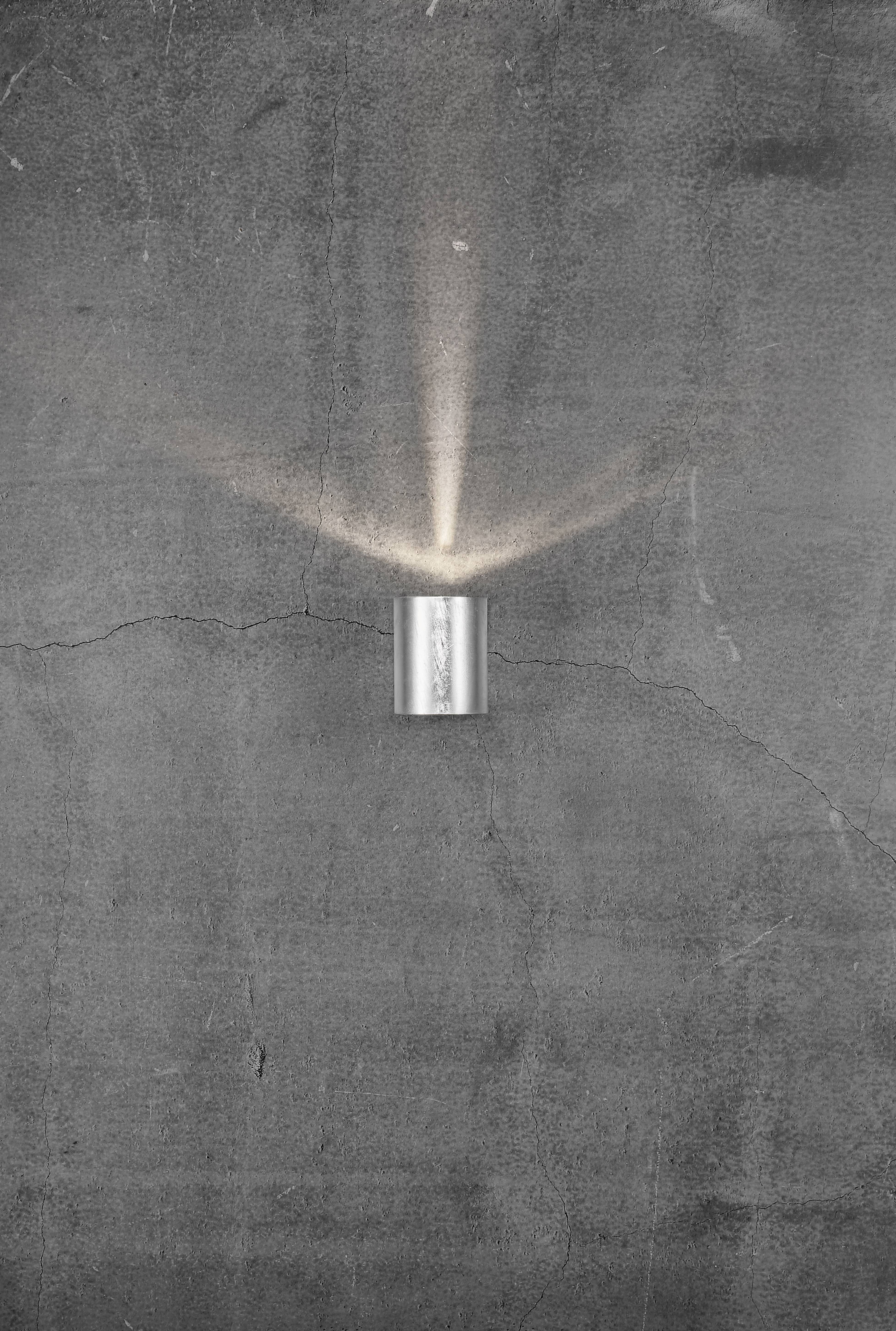 Eine silberne Wandlampe leuchtet nach oben und unten auf eine graue Betonwand mit sichtbaren Rissen.