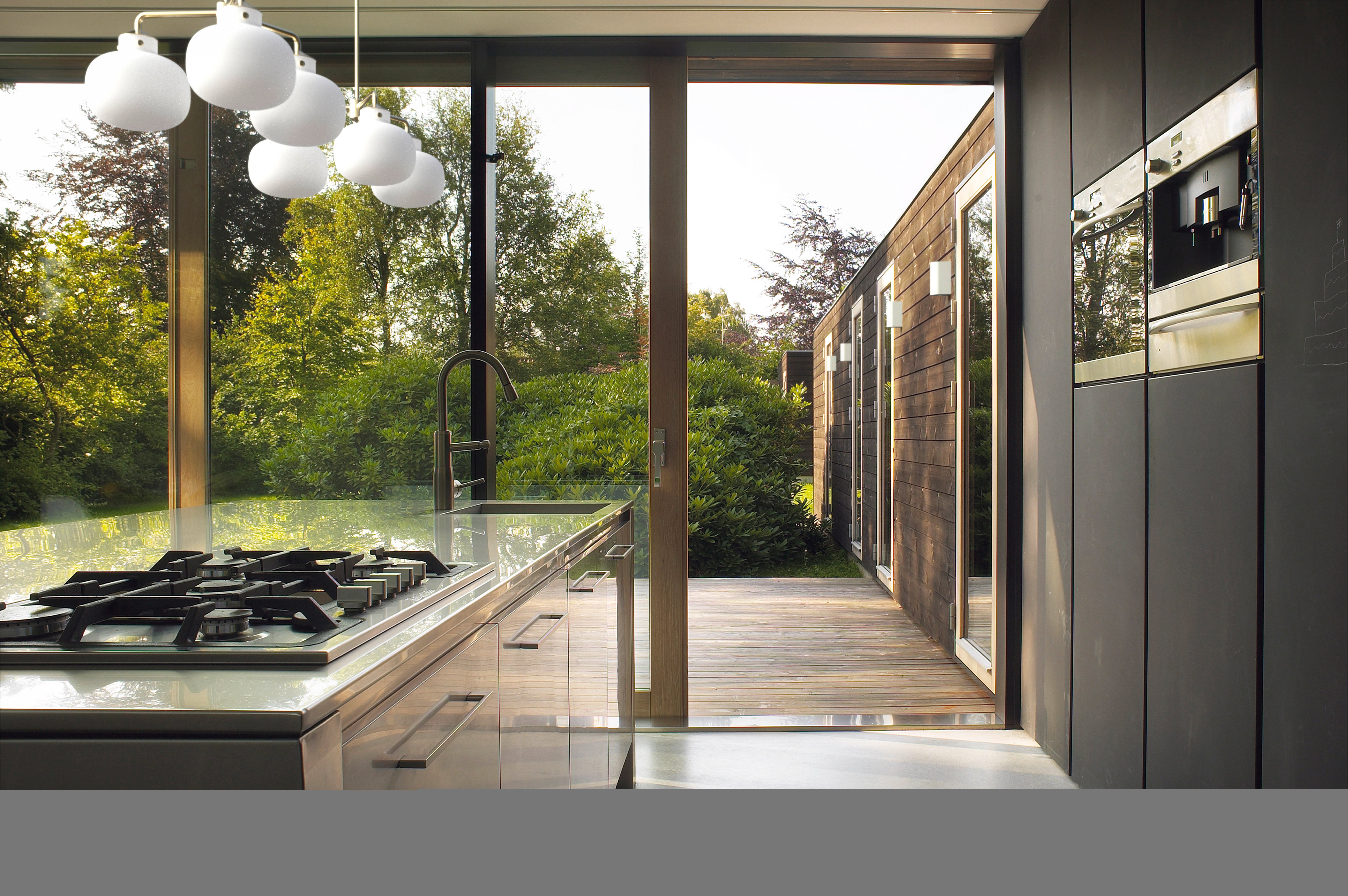 Moderne Küche mit großem Fenster, das auf einen grünen Garten blickt. Glatte Oberflächen, helle Beleuchtung, offenes Design für nahtlosen Übergang nach draußen.
