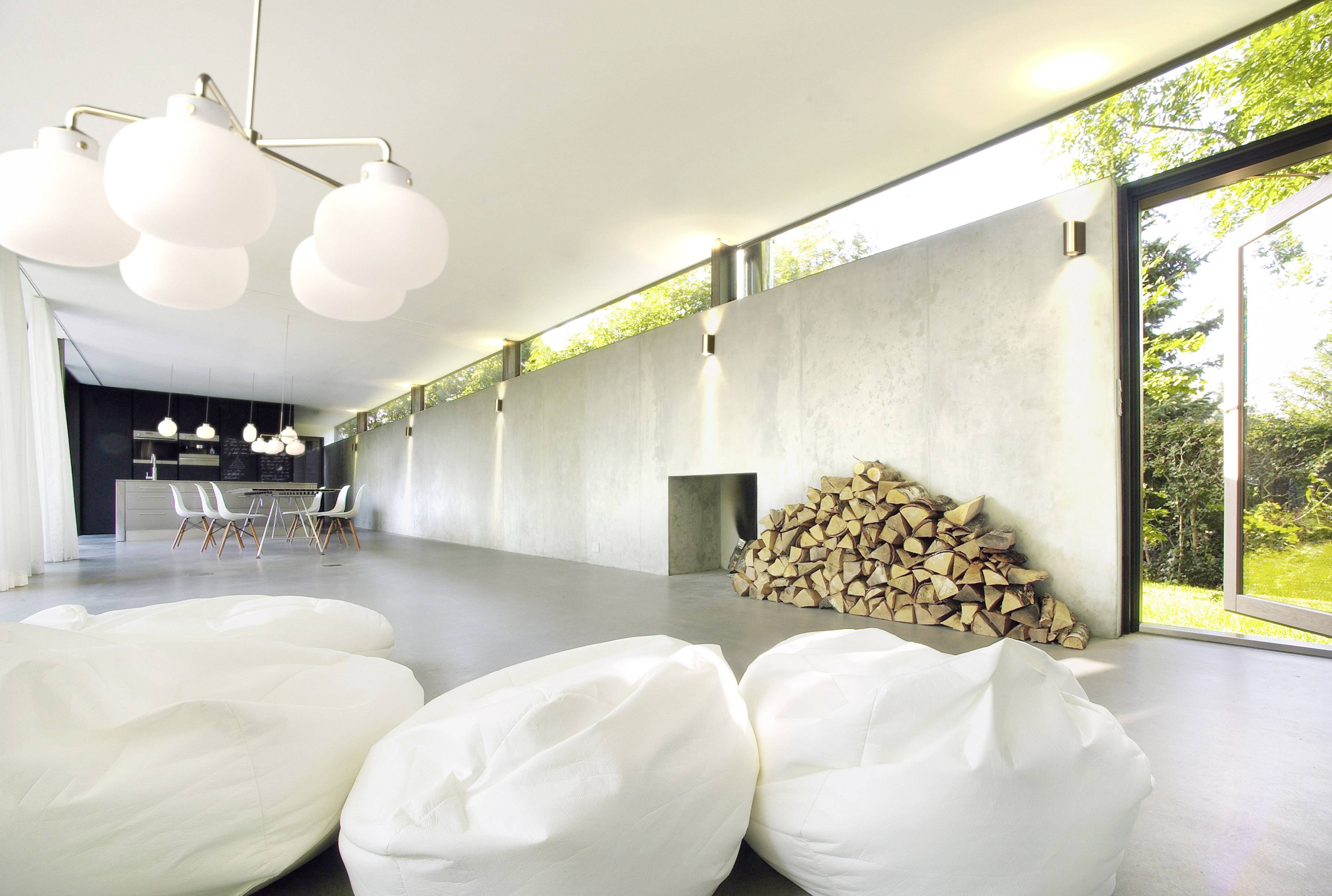 Moderner Wohnraum mit großen Fenstern, weißen Sitzsäcken und einem Stapel Holz an der Wand. Helle und minimalistische Einrichtung.