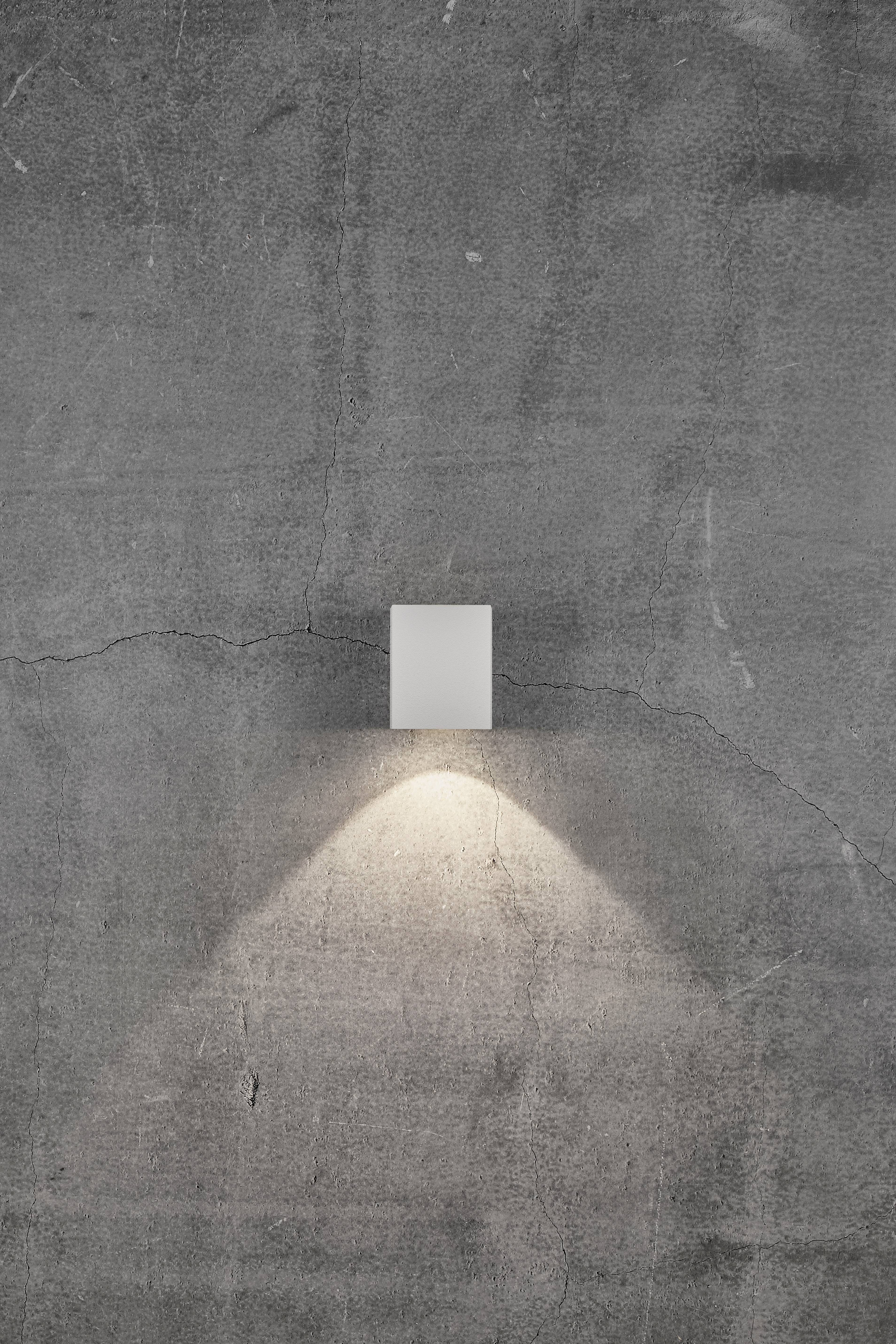 Eine Wandlampe leuchtet auf eine graue, rissige Betonwand und erzeugt ein dreieckiges Lichtmuster nach unten.