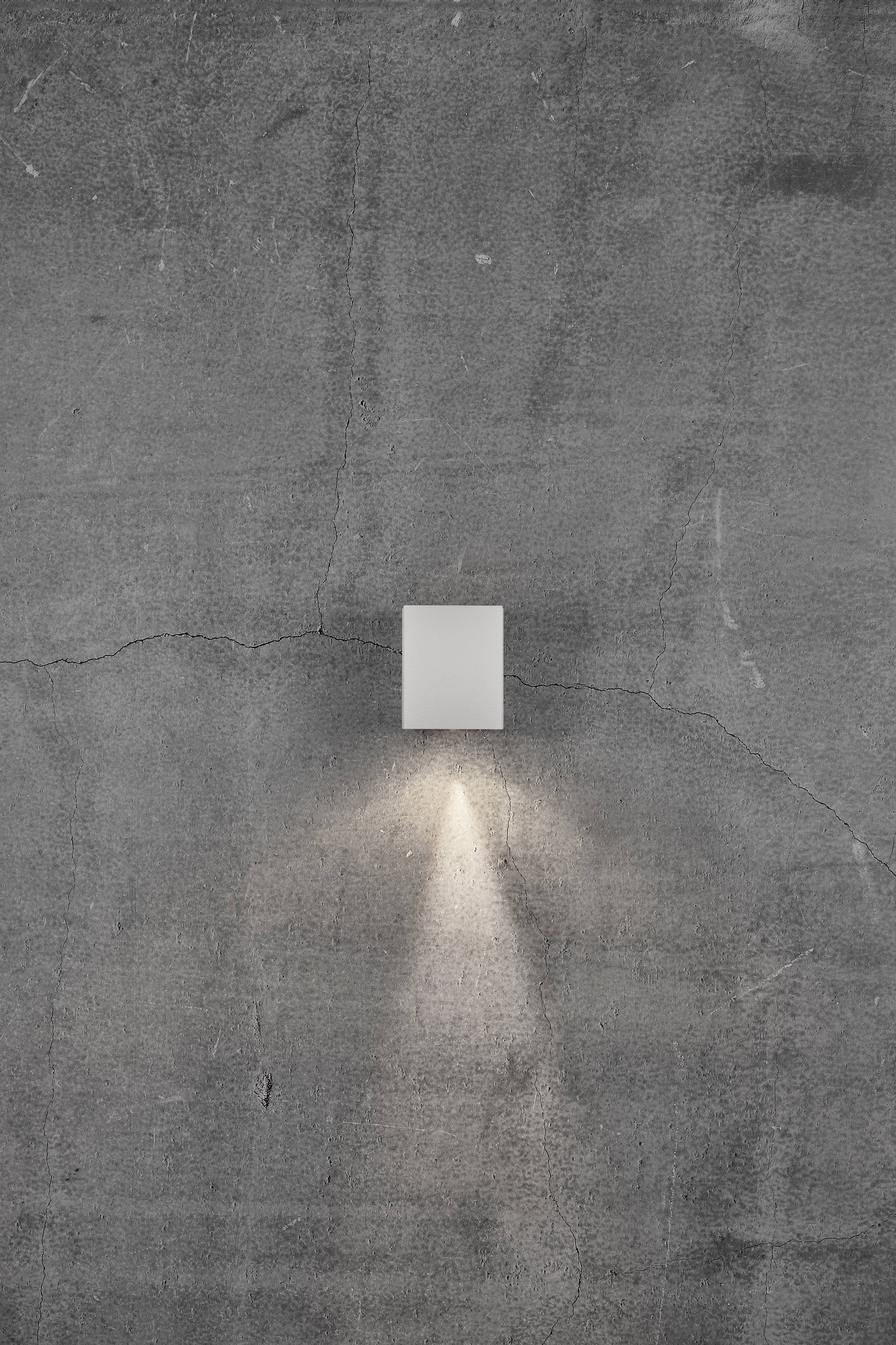 Eine kleine, rechteckige Wandlampe an einer grauen, rissigen Betonwand. Die Lampe strahlt ein schmales, nach unten gerichtetes Licht aus.