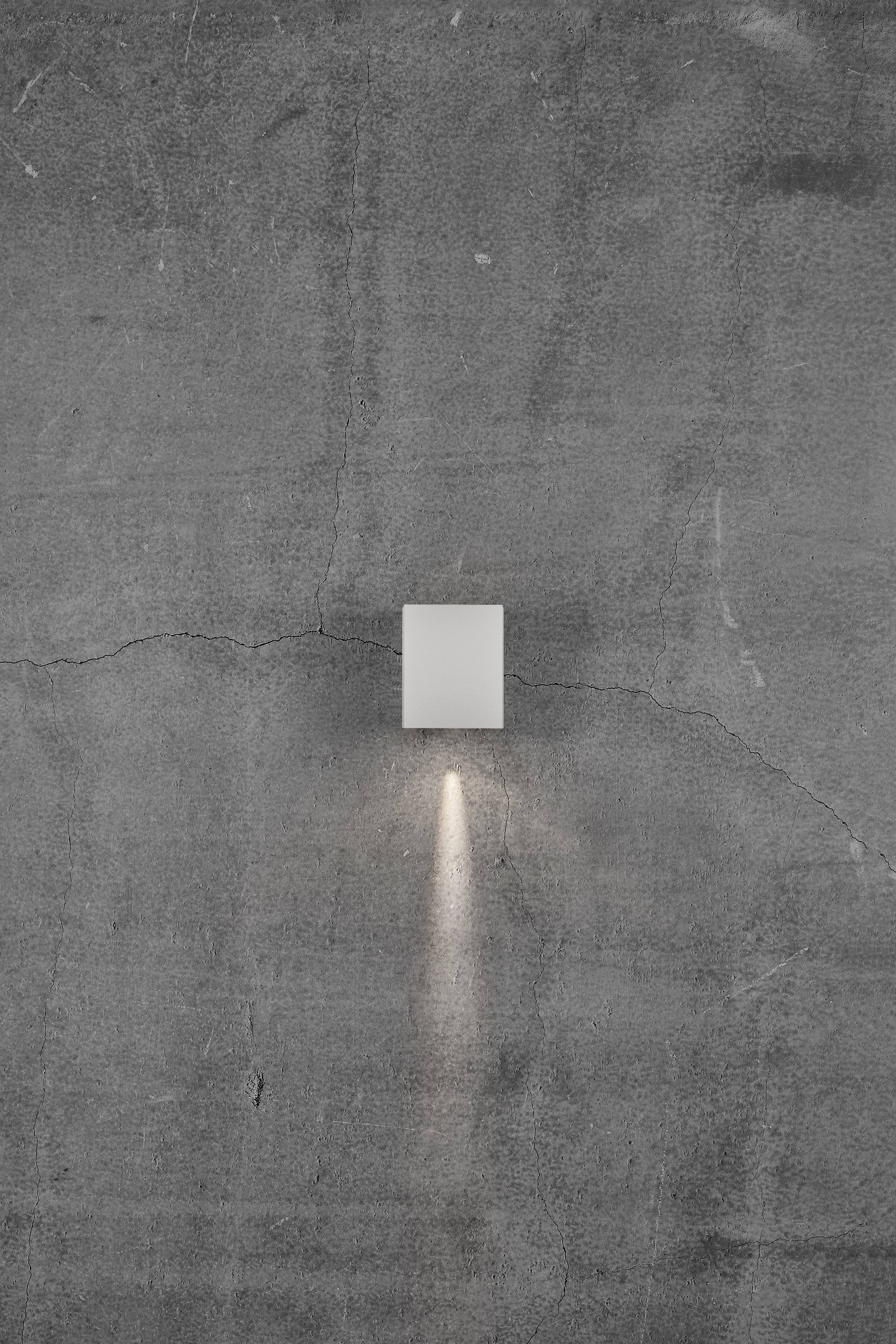 Wandleuchte an einer grauen Wand, wirft einen Lichtstrahl nach unten und erzeugt so einen warmen, modernen Lichteffekt.