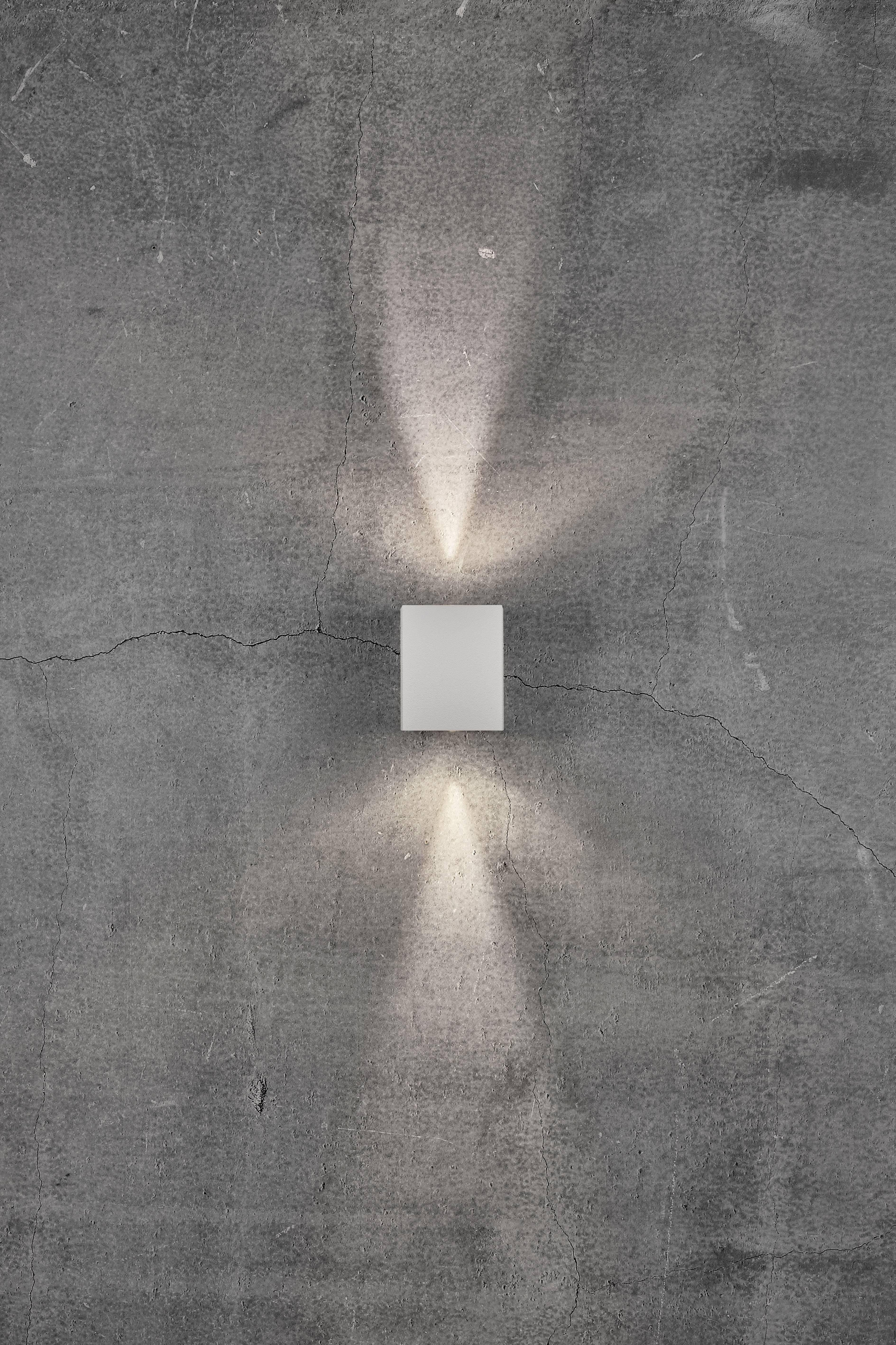 Eine moderne Wandleuchte auf grauer, leicht rissiger Betonwand. Licht strahlt nach oben und unten, erzeugt symmetrische Muster.