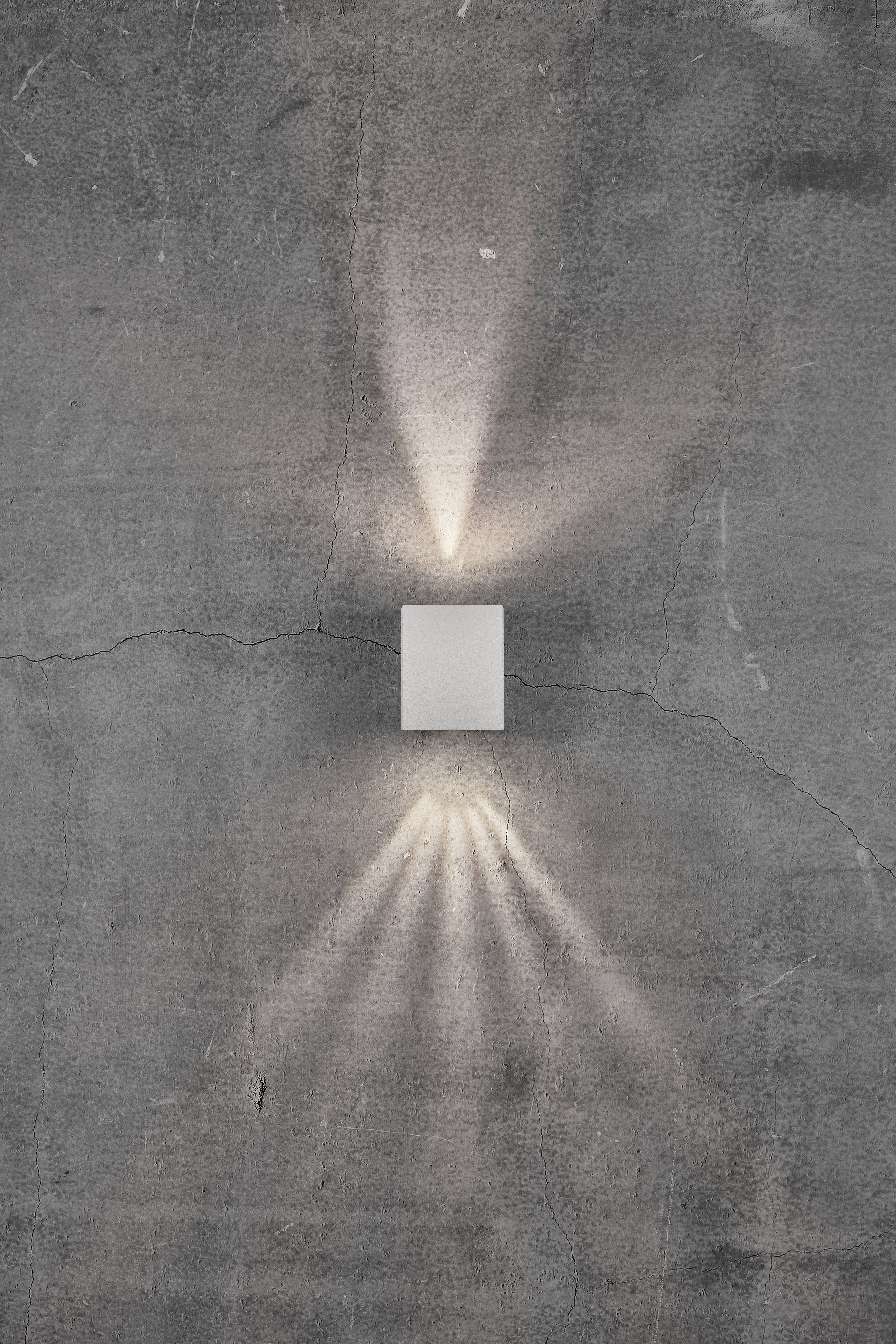 Eine Wandleuchte auf rauem Beton, wirft Lichtstrahlen nach oben und unten, was ein dekoratives Muster bildet.