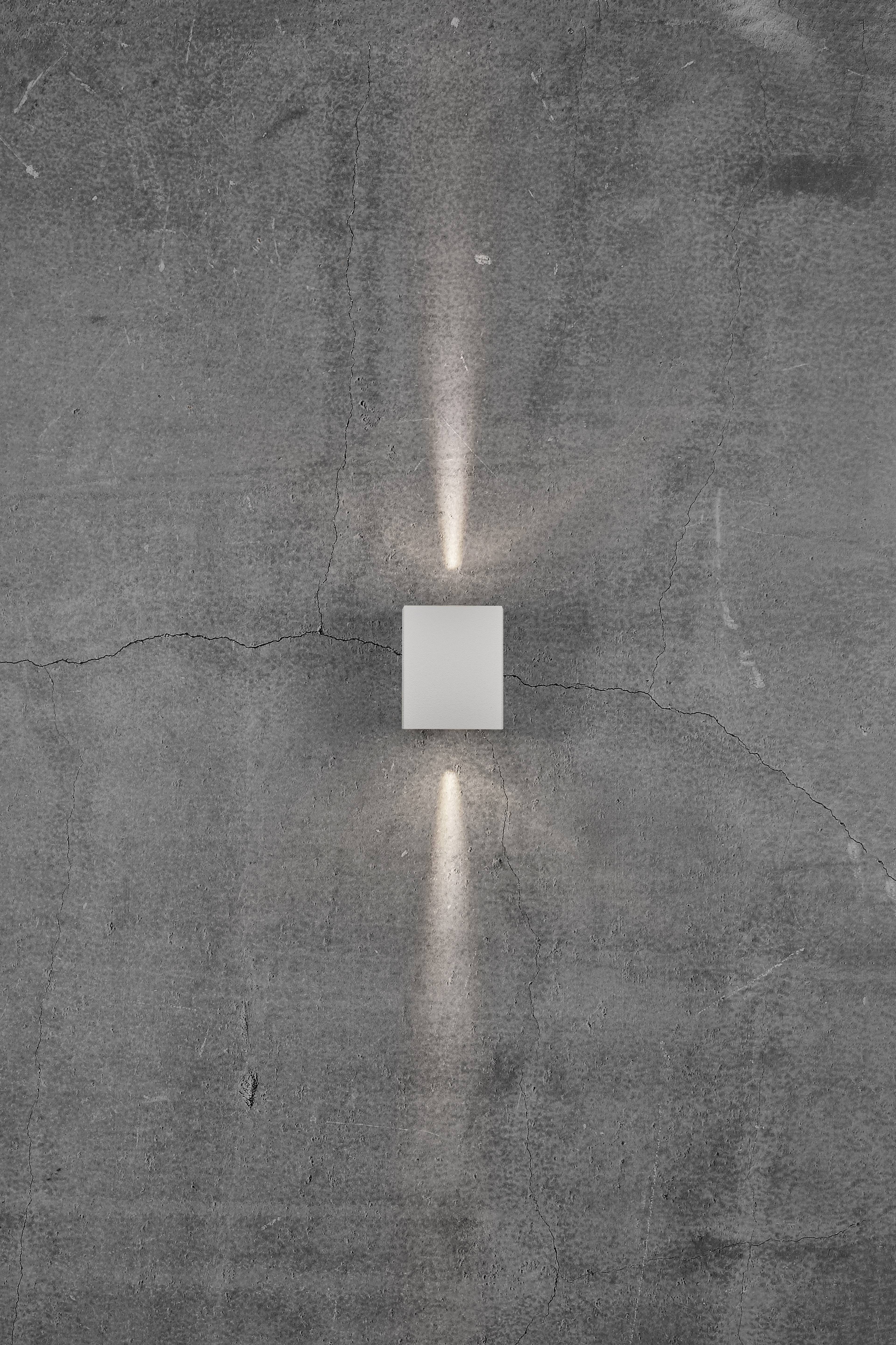 Eine schlichte weiße Wandleuchte strahlt Licht nach oben und unten auf eine graue Betonwand.