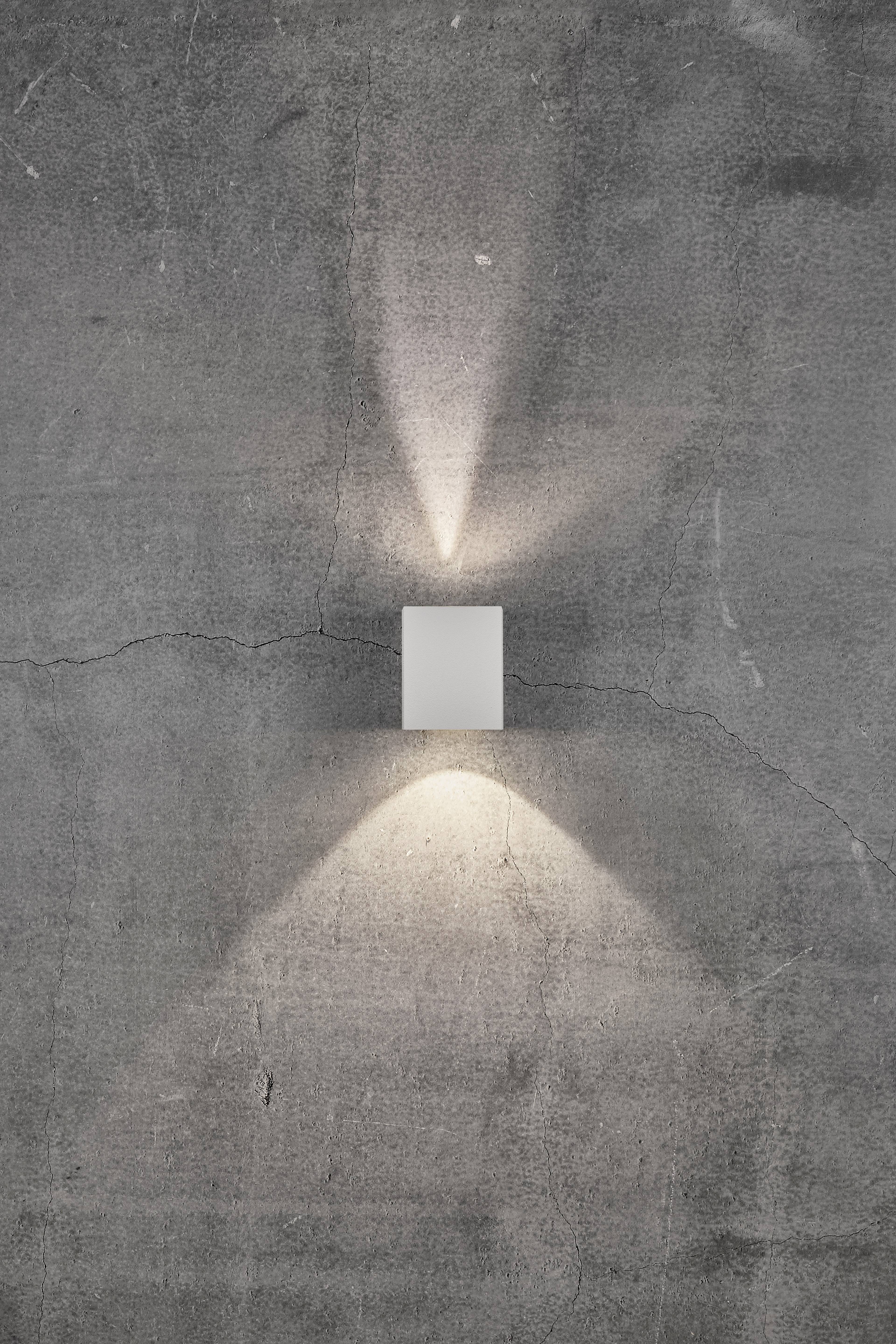 Ein weißer Wandstrahler an einer grauen Betonwand, strahlt Licht nach oben und unten und erzeugt ein geometrisches Muster.