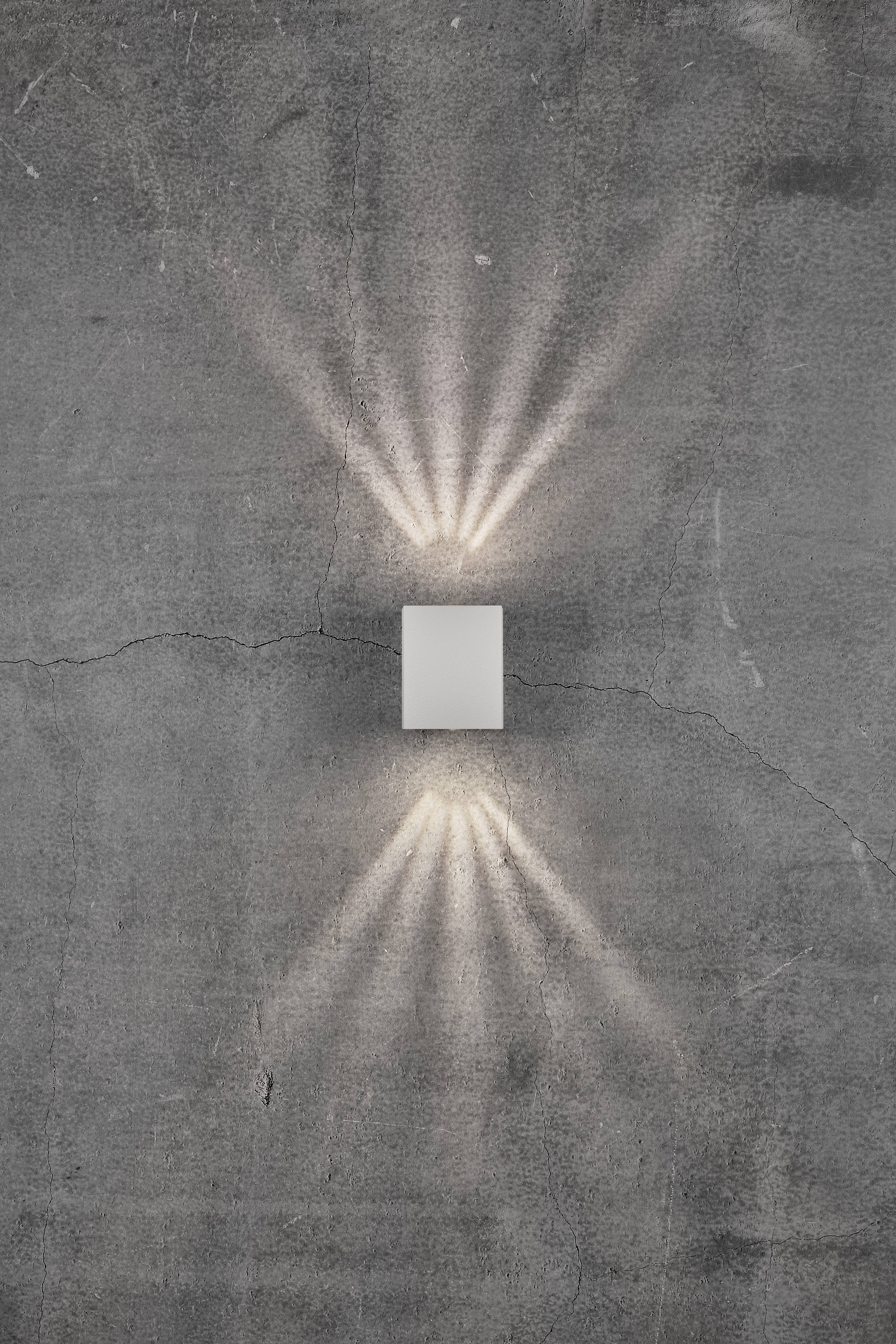 Eine Wandleuchte an einer grauen Wand, deren Lichtstrahlen fächerartig nach oben und unten leuchten.