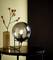 Runde Tischlampe mit rauchgrauem Glas und schwarzem Gestell auf einem Tisch. Daneben steht eine Vase mit Zweigen vor dunklem Hintergrund.