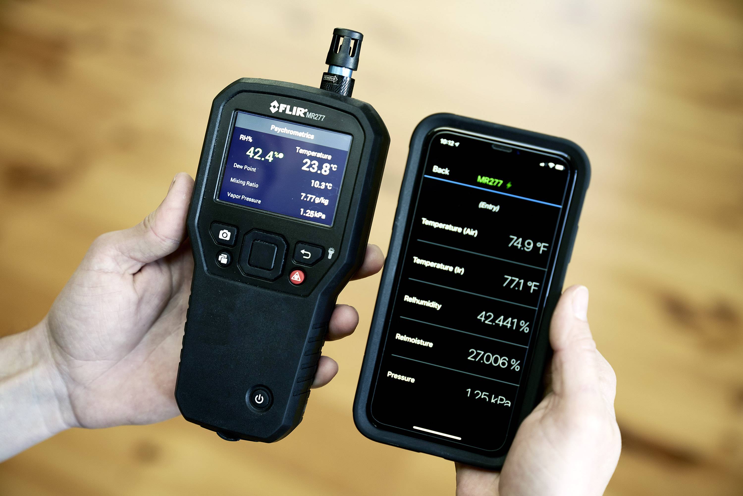 Eine Person hält ein Handgerät und ein Smartphone. Das Handgerät zeigt CO2-Werte und Temperatur an, während das Smartphone ähnliche Messdaten anzeigt.