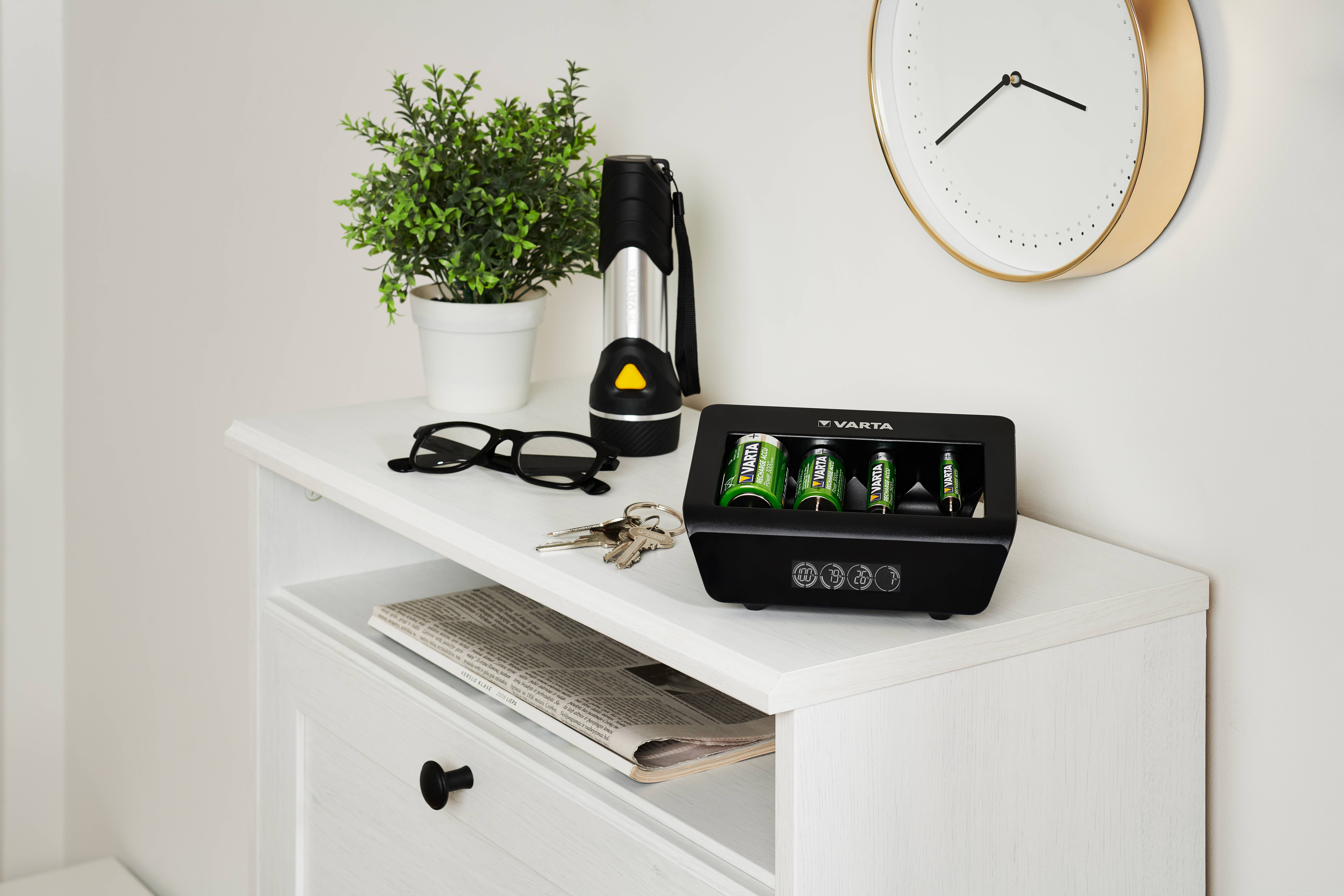 Ein kleiner Tisch mit einer Pflanze, einer Taschenlampe, einer Brille, Schlüsseln und einem Varta-Ladegerät mit Batterien. Eine Uhr hängt an der Wand darüber.