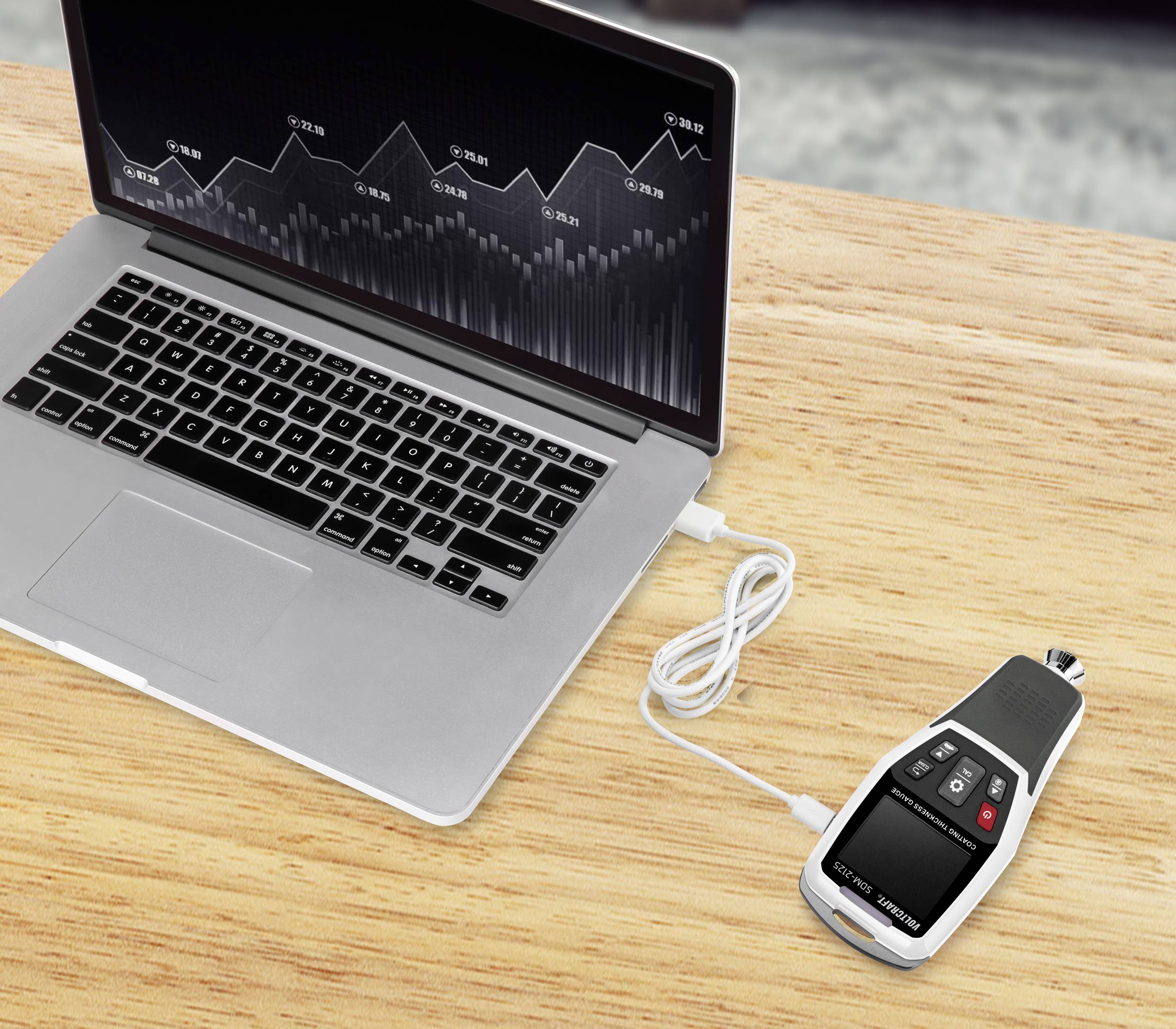 Ein Laptop mit einer grafischen Analyse auf dem Bildschirm ist per USB-Kabel mit einem Messgerät verbunden, das auf einem Holztisch liegt.