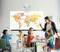 Eine Lehrerin steht vor einer Weltkarte an der Tafel und unterrichtet Kinder, die an Tischen sitzen. Arrows zeigen Richtung Nordamerika.