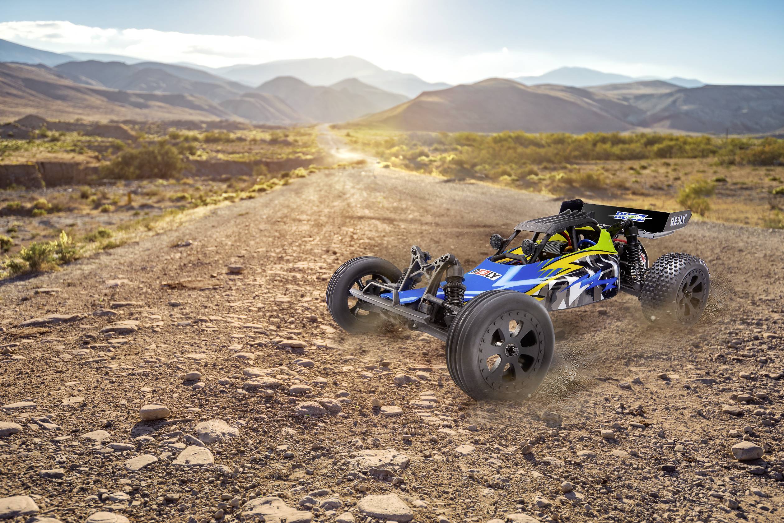 Ein ferngesteuertes Auto fährt auf einer sandigen, steinigen Straße in einer wüstenartigen Landschaft. Berge sind im Hintergrund sichtbar.