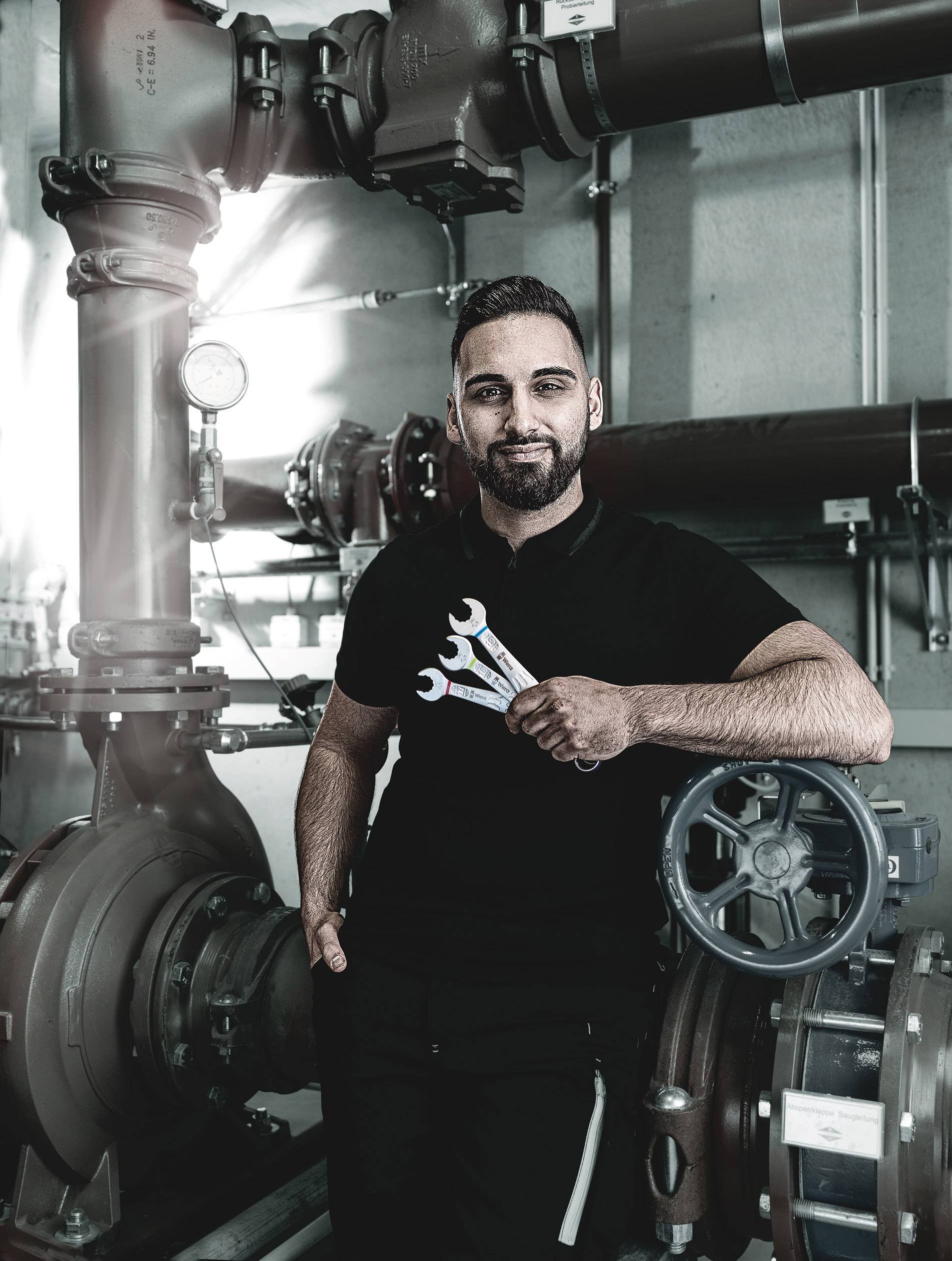 Ein Mann hält Werkzeuge in einem technischen Raum mit großen Rohren und Ventilen, möglicherweise ein Wartungstechniker bei der Arbeit.