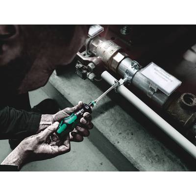 Eine Person zieht eine Schraube an einem Rohrsystem mit einem grünen Schraubendreher an, in einem industriellen Umfeld.
