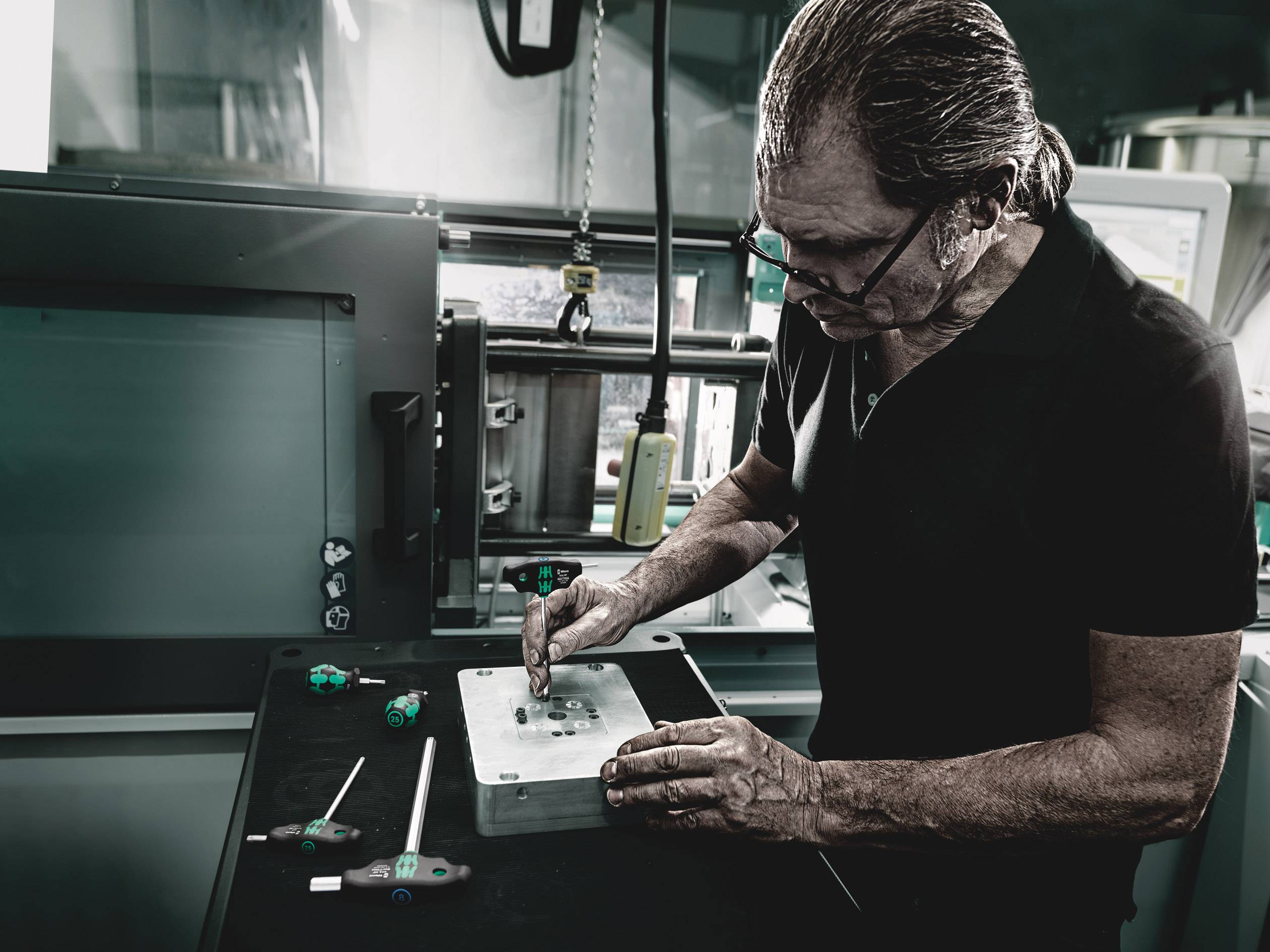 Eine Person arbeitet an einem Metallwerkstück in einer Werkstattumgebung, umgeben von Werkzeugen und Maschinen.