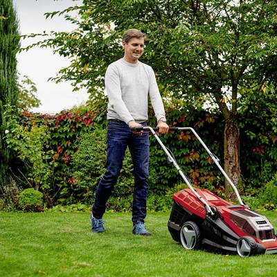 Ein Mann mäht den Rasen in einem Garten. Er trägt einen grauen Pullover und blaue Schuhe. Im Hintergrund sind Bäume und Sträucher zu sehen.