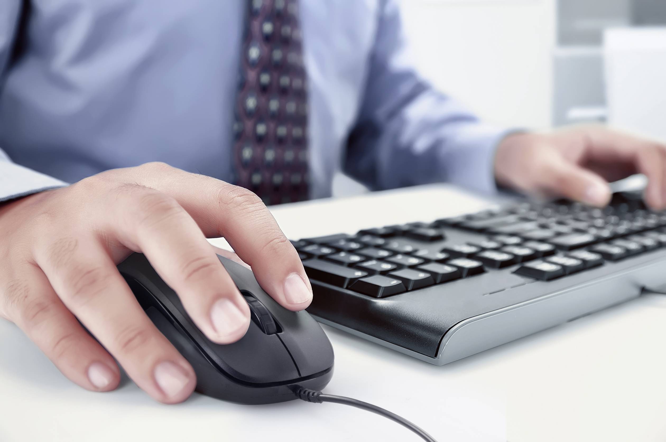 Eine Person sitzt an einem Schreibtisch und benutzt eine Computermaus und eine Tastatur. Die Person trägt ein Hemd und eine Krawatte.