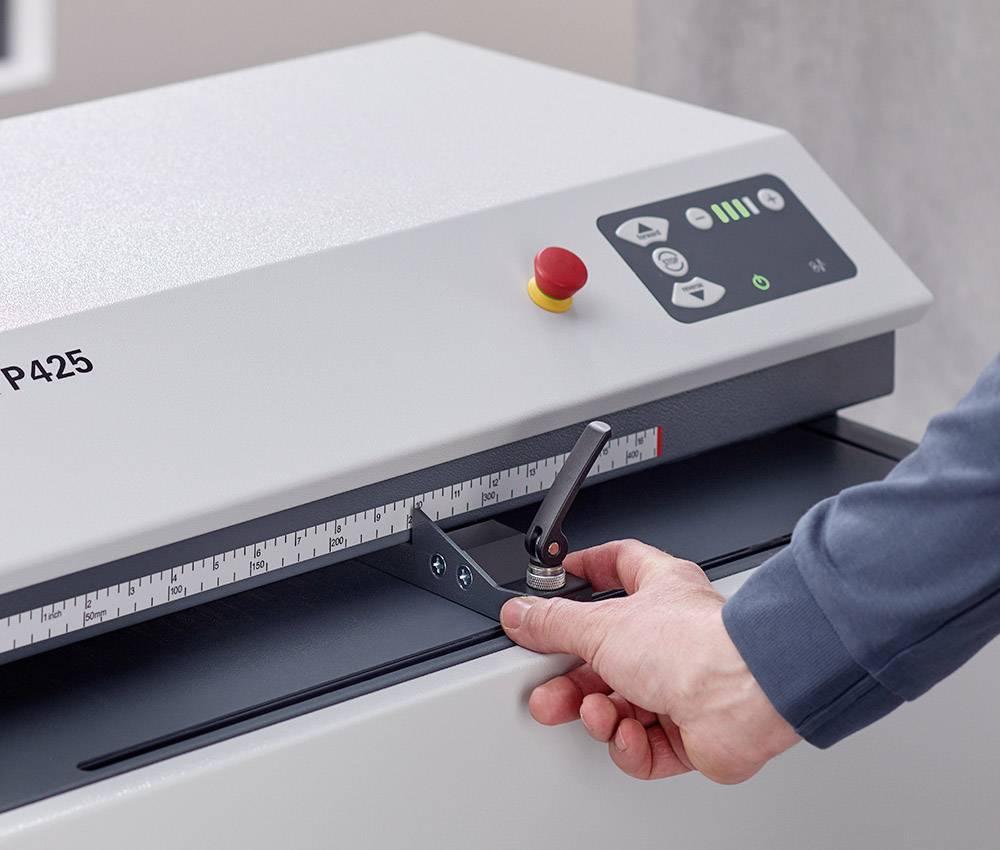 Eine Person bedient eine industrielle Papiermaschine, indem sie den Hebel justiert. Die Maschine zeigt eine Skala zur Längeneinstellung an.