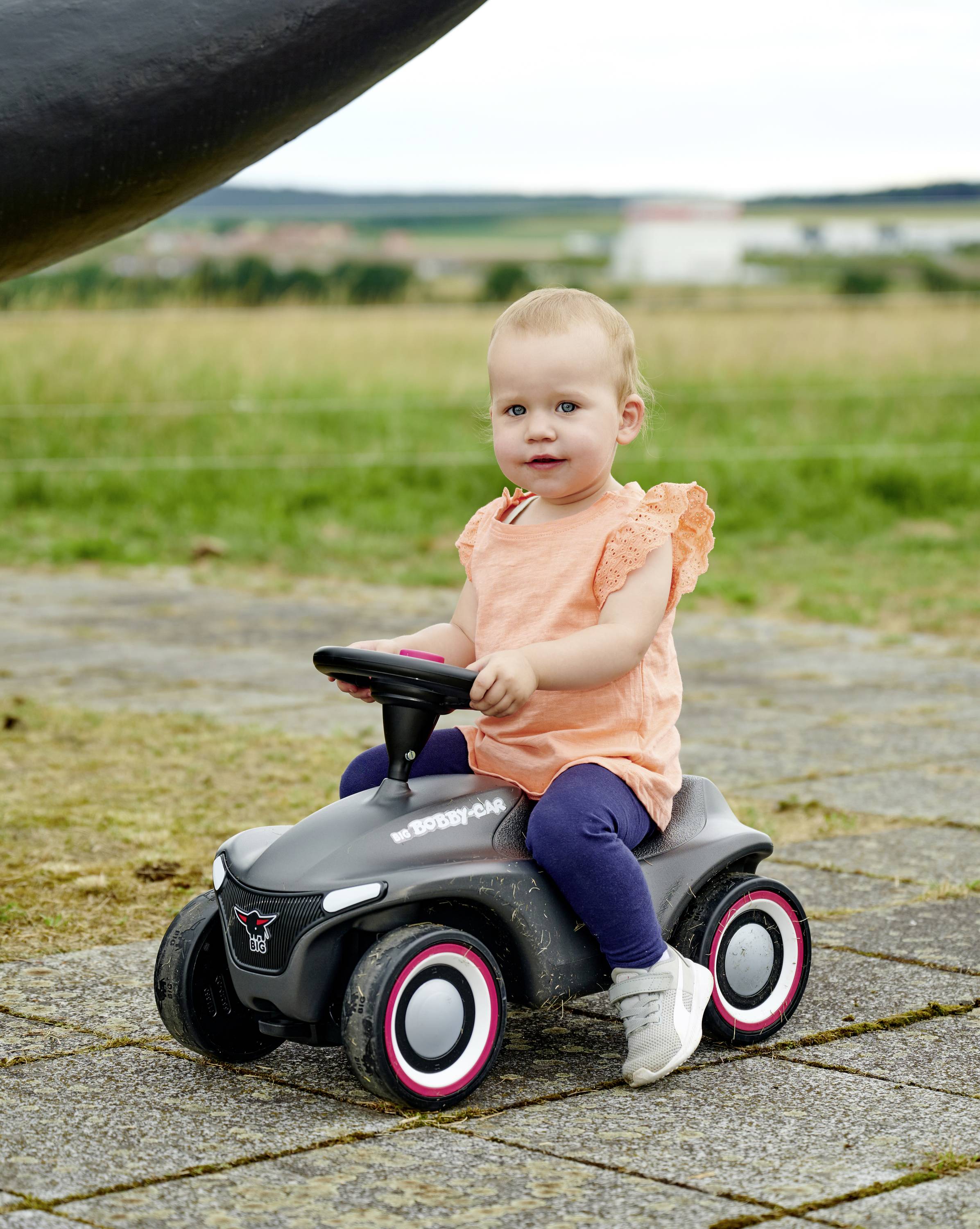 Kleinkind sitzt auf einem schwarzen Rutschauto im Freien, trägt ein orangefarbenes Oberteil und blaue Hosen, mit grünem Feldhintergrund.