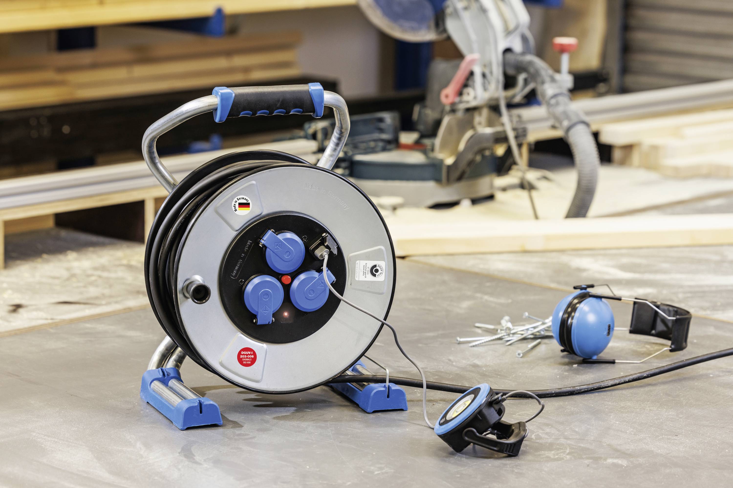 Eine Kabeltrommel liegt auf einer Werkbank in einer Werkstatt mit Holzarbeiten im Hintergrund, neben einem Maßband und Baugegenständen.